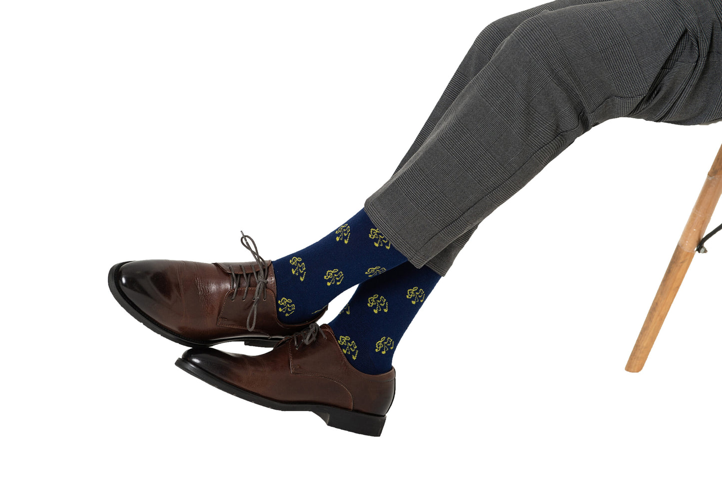 A person sits cross-legged on a chair, wearing gray plaid pants, brown dress shoes, and blue Musical Note Socks with yellow patterns—a stylish look that’s perfect for music lovers.