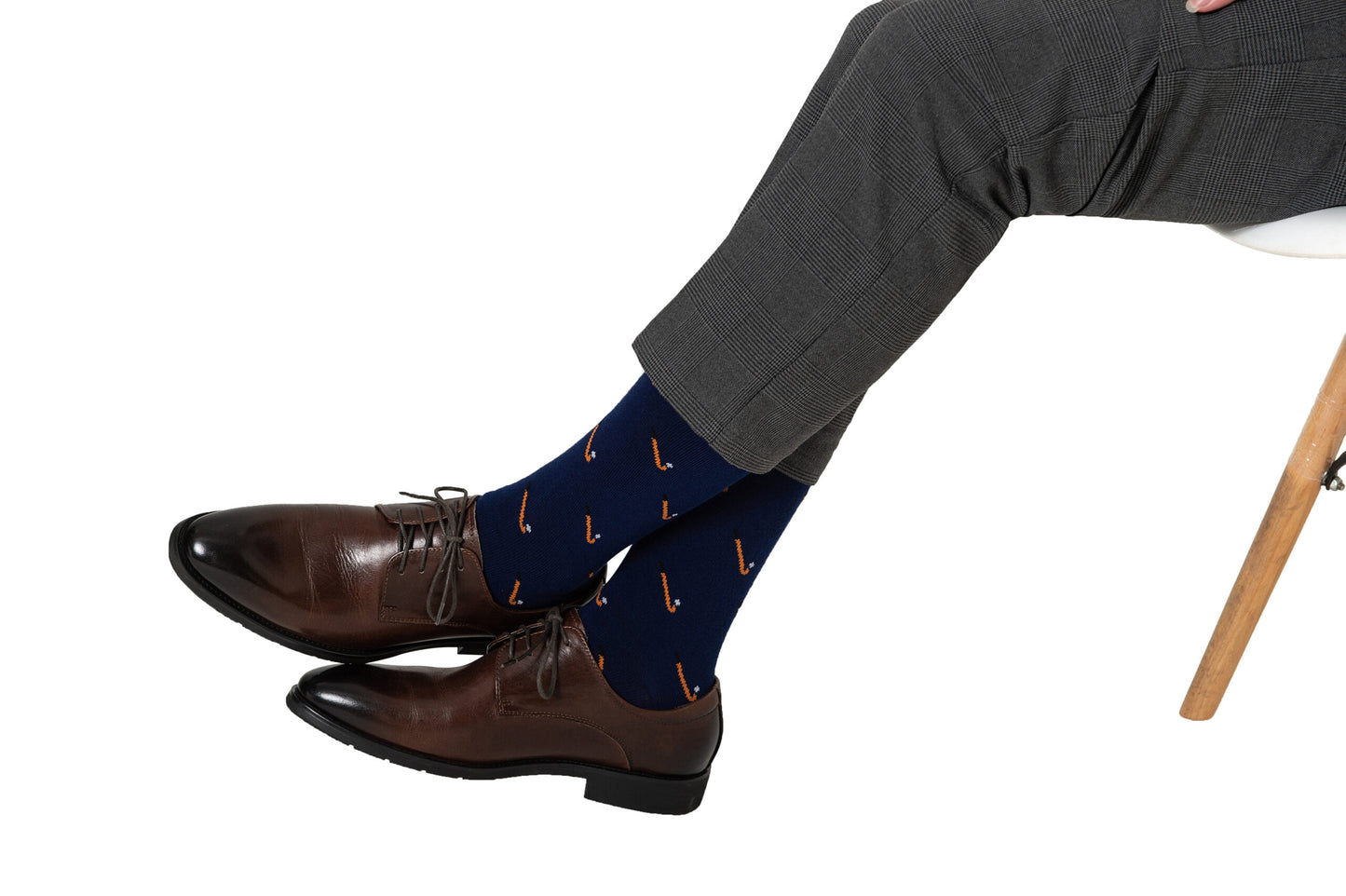 A person sits cross-legged on a white chair, wearing gray pants, brown dress shoes, and Field Hockey Socks—navy moisture-wicking socks with a red pattern.