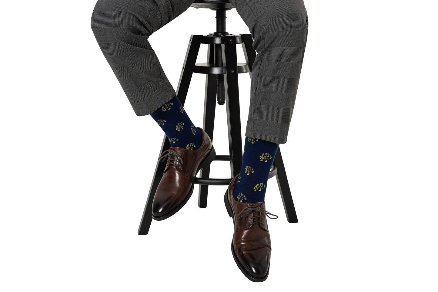 Wearing gray dress pants and brown shoes, a person sits on a black stool to display blue Musical Note Socks with a yellow pattern—an ideal accessory for musicians and music lovers.
