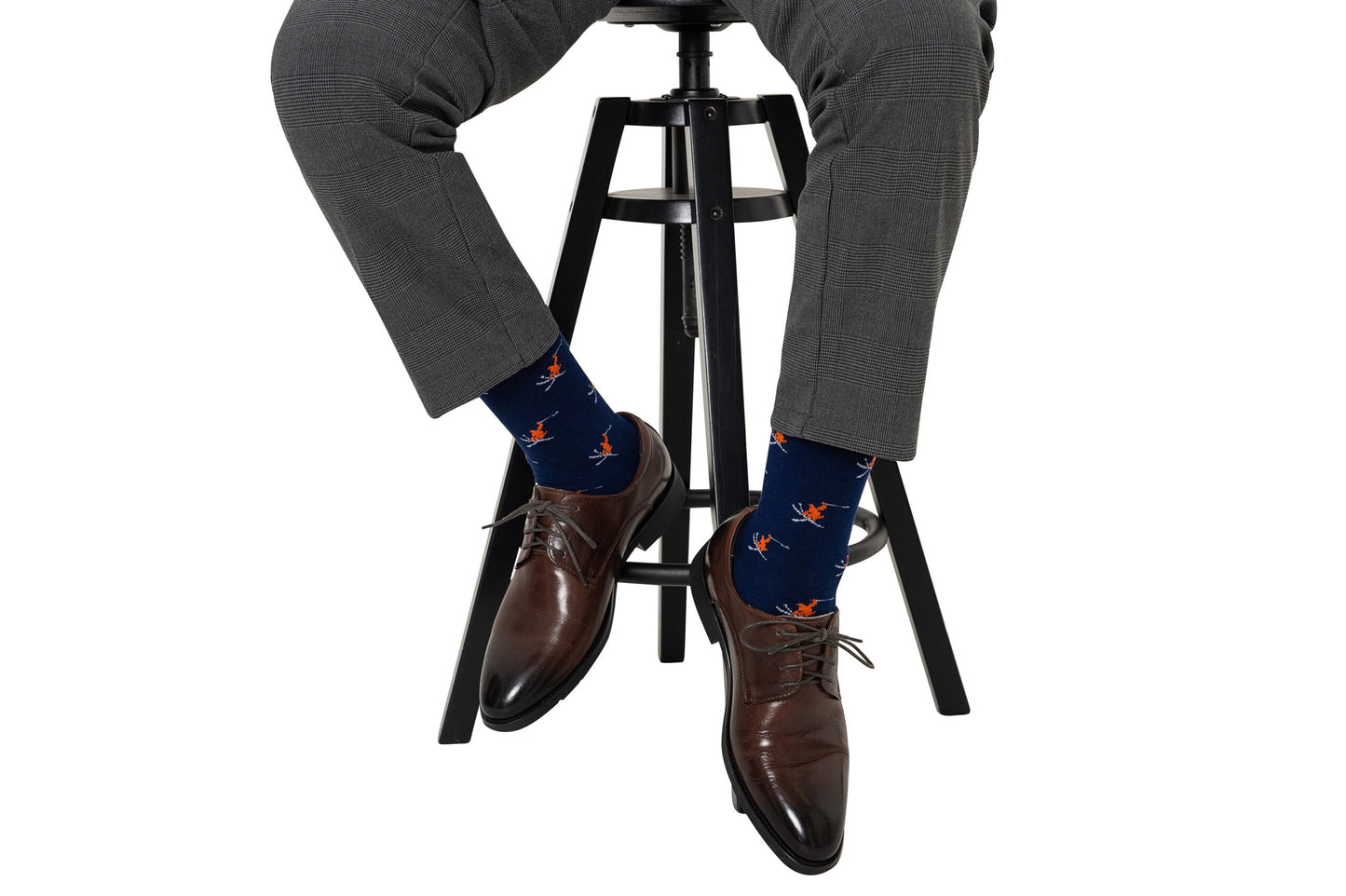 A person in gray plaid pants and brown dress shoes sits on a black stool, wearing Ski Socks in navy blue with an orange bird pattern.