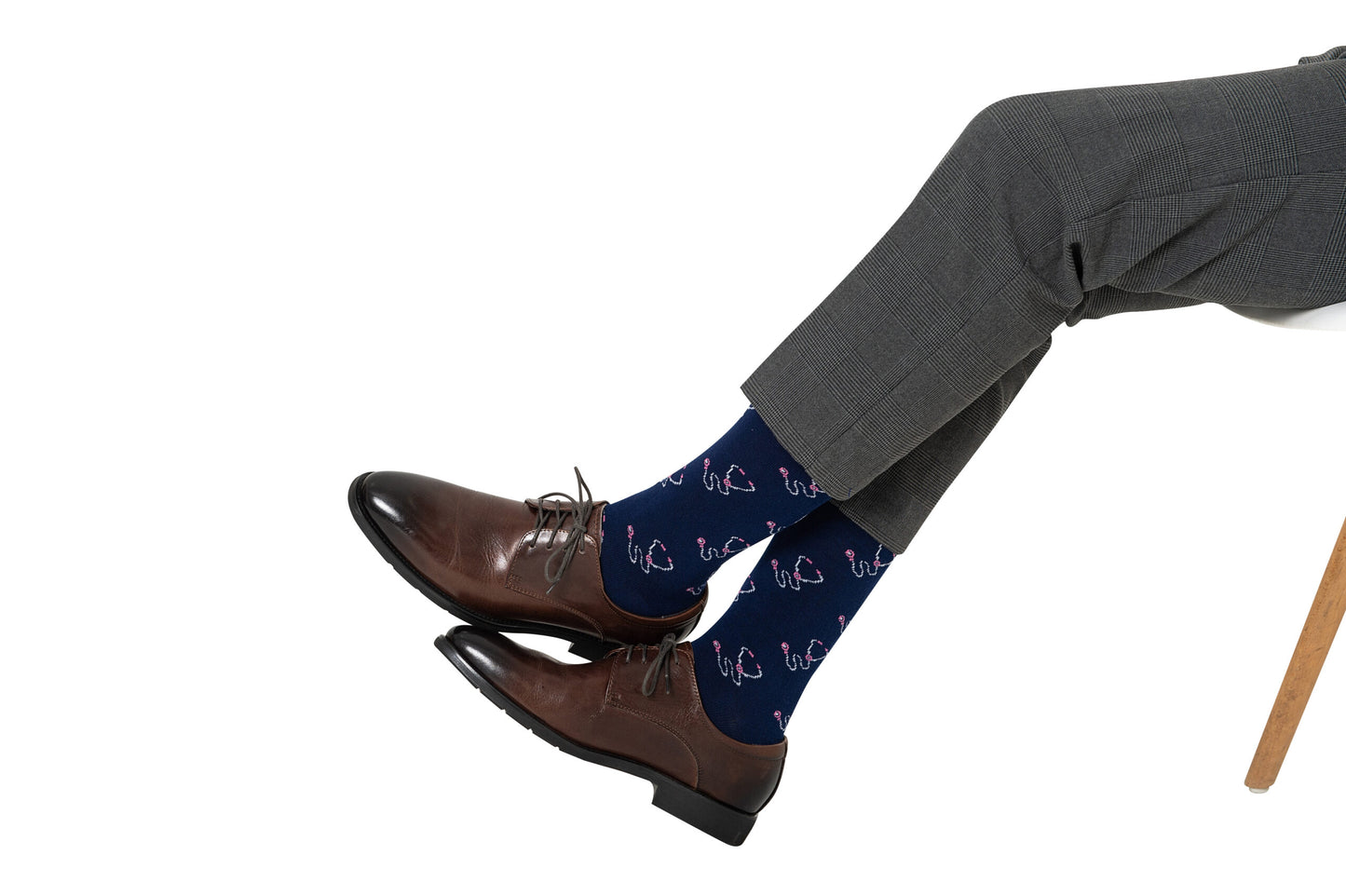 A person wearing dark dress pants, Stethoscope Socks featuring a pink flamingo pattern, and brown dress shoes sits on a white chair.