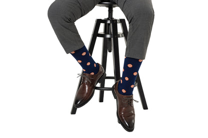 A person seated on a stool wearing gray trousers, orange socks with citrusy comfort orange circle patterns, and brown dress shoes.