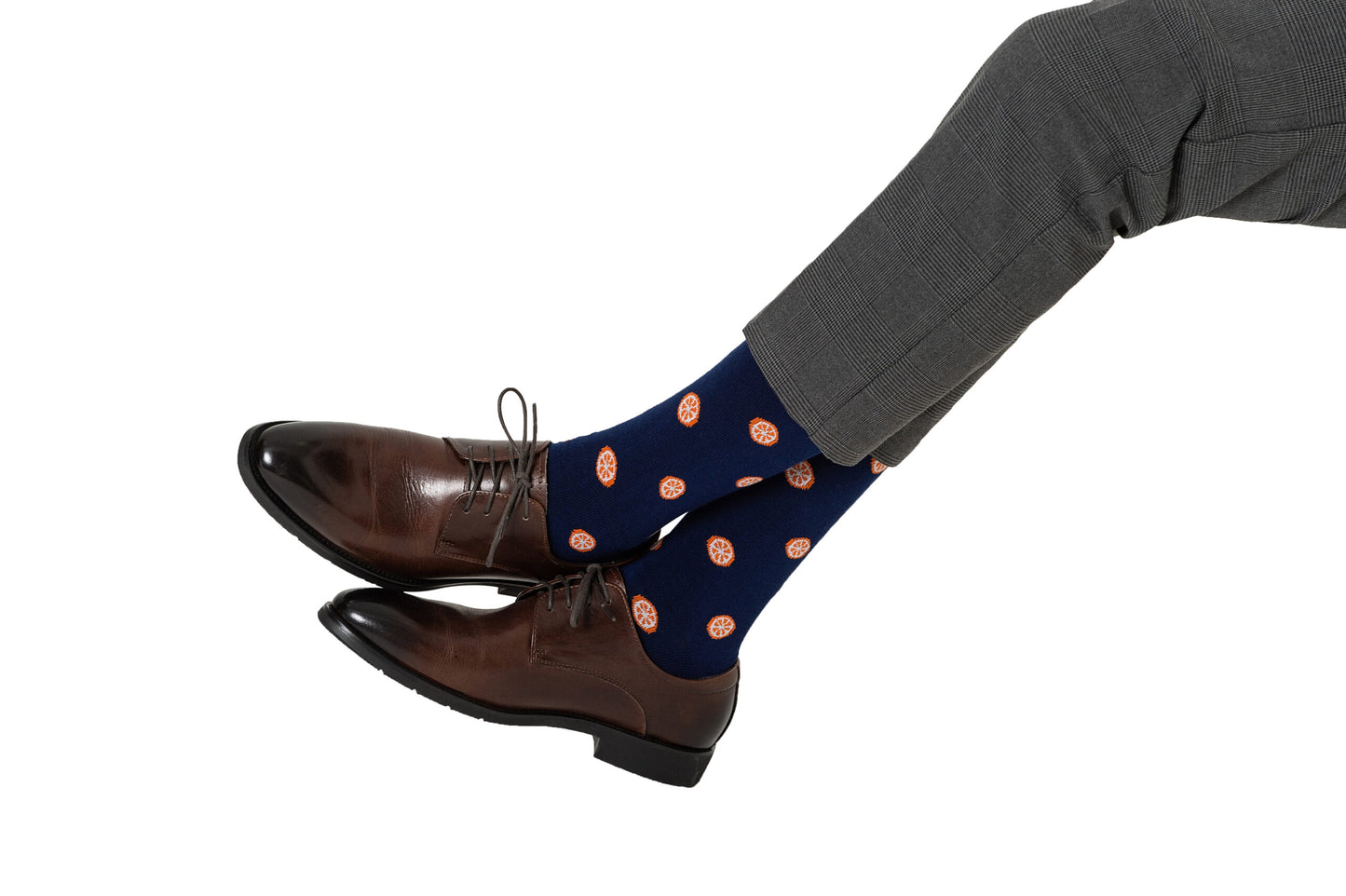 A person wearing brown leather shoes and grey trousers with Blue Socks adorned with citrusy Orange patterns, crossed at the ankles heel to toe against a white background.