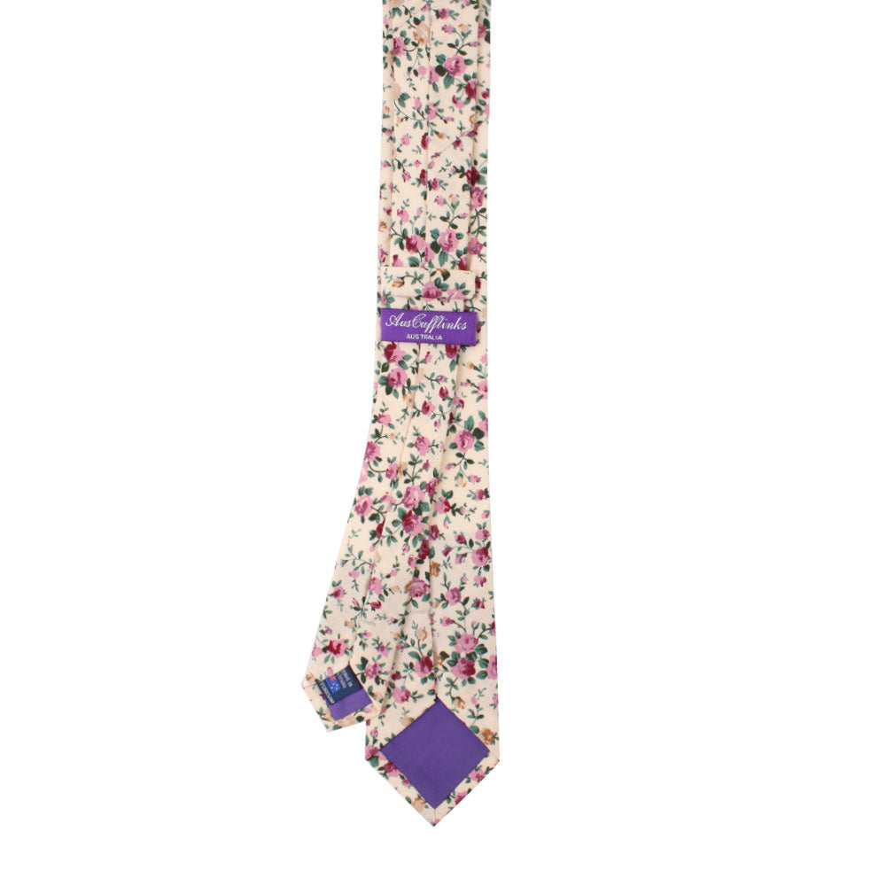 A Pastel Pink Rose Floral Cotton Skinny Tie, elegantly featuring a cream-colored base adorned with pink and green rose patterns, complete with its signature purple label and tastefully detailed ends.