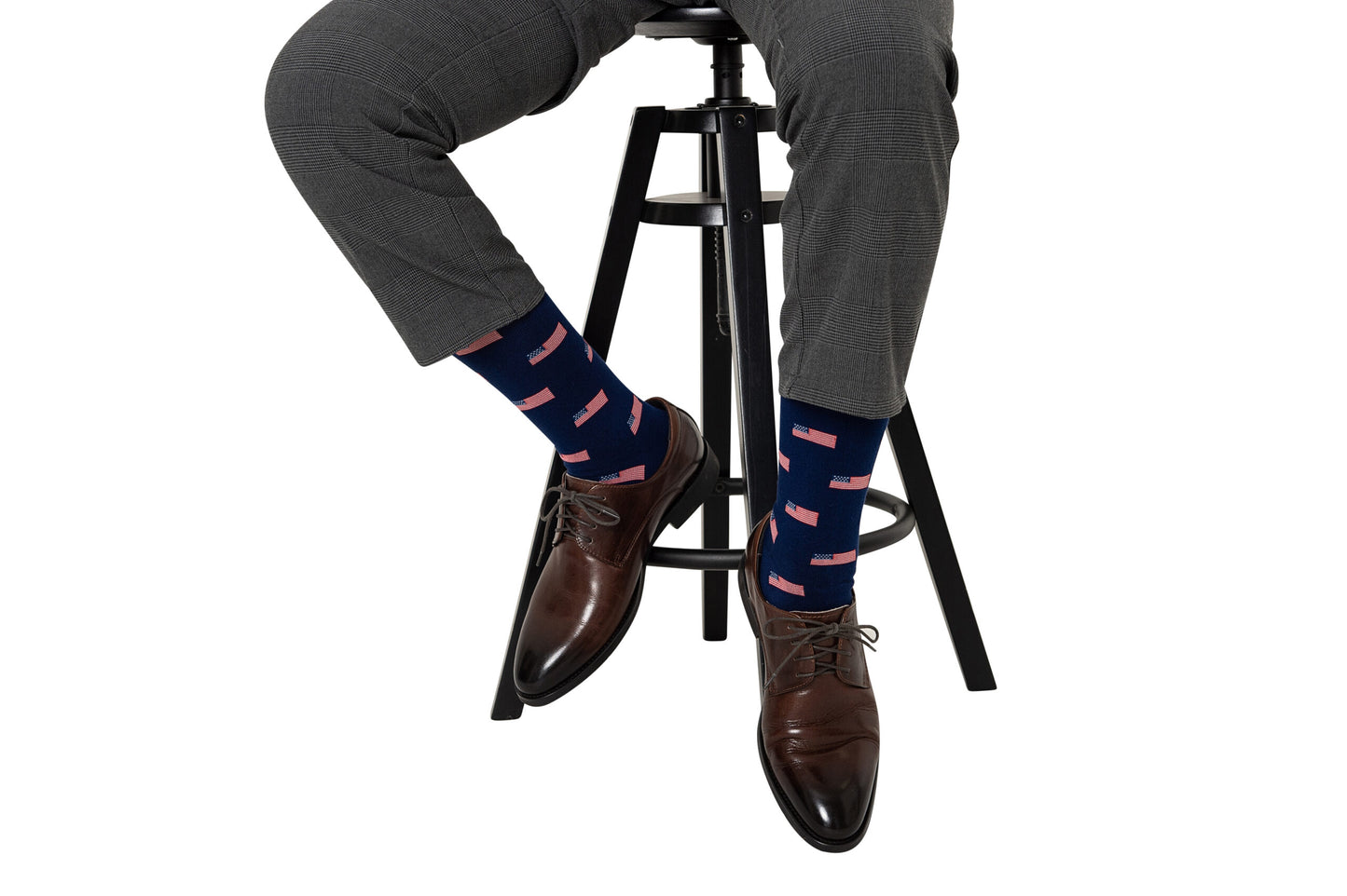 A person sits on a black stool, wearing gray pants and brown shoes, adding a touch of style with USA Flag Socks that feature stars and stripes.