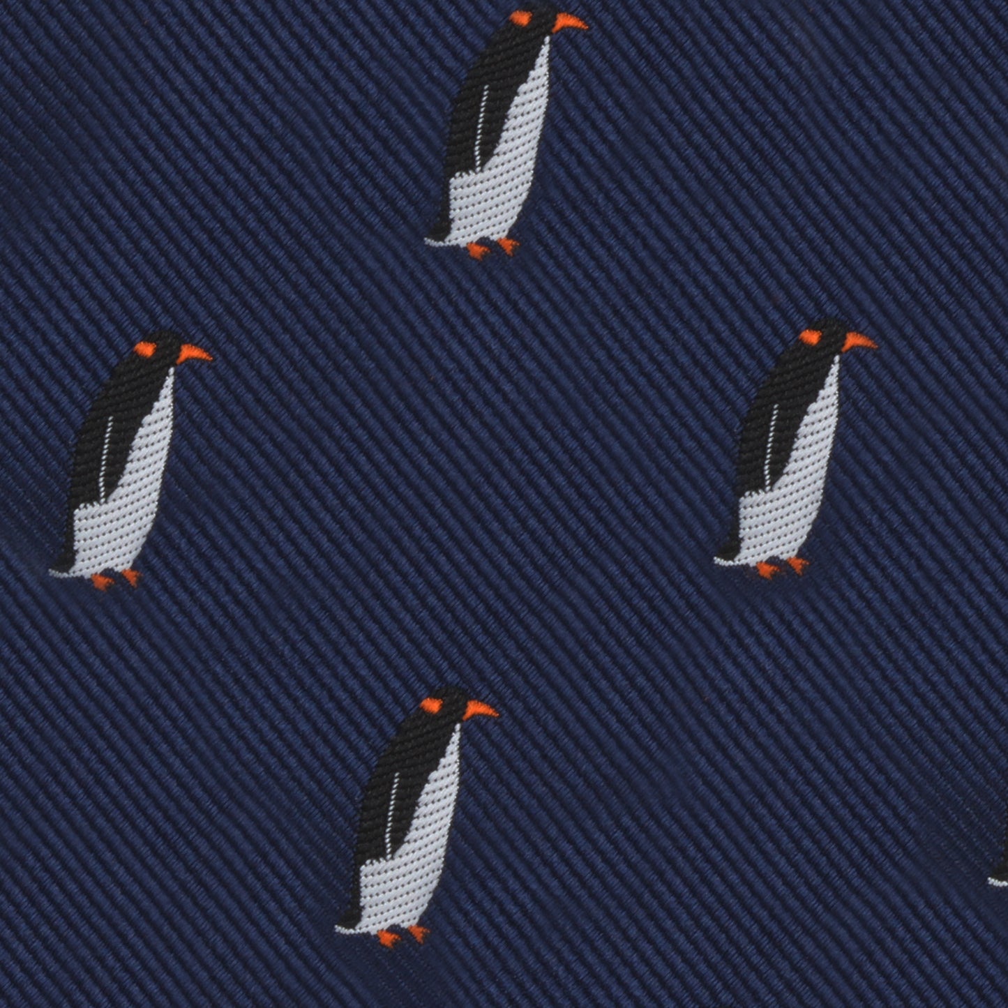 Close-up of a dark blue fabric with an embroidered pattern of penguins with orange beaks and feet, evenly spaced throughout the Penguin Bow Tie, exuding a cold elegance that contrasts beautifully with its charming design.