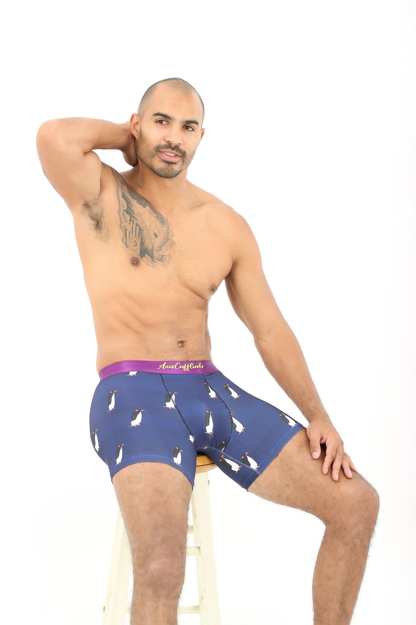 A man with a tattoo on his chest sits on a stool, relaxing in Penguin Underwear adorned with purple penguin patterns, embodying polar comfort.