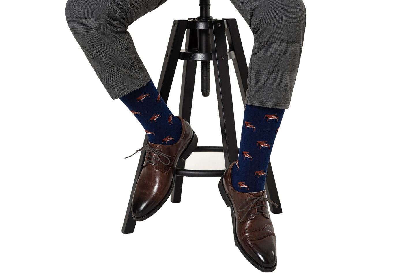 Someone wearing gray pants, brown dress shoes, and Piano Socks with a navy blue background and fox pattern sits on a black stool—an ideal choice for unique musician gifts.