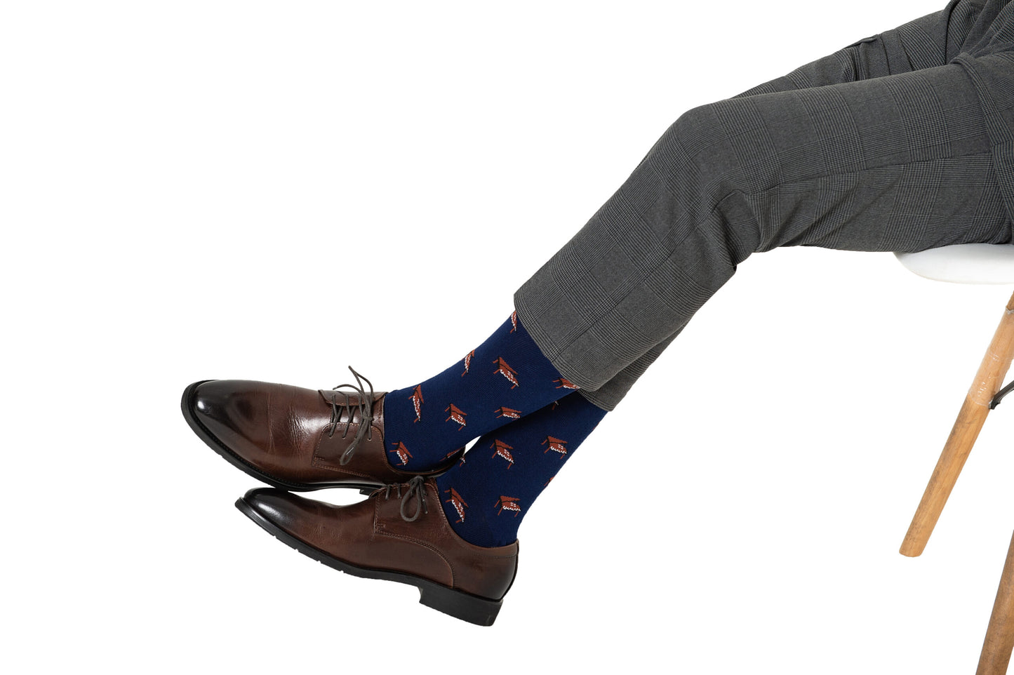 A person wearing gray dress pants, brown dress shoes, and Piano Socks in blue sits on a white chair with legs crossed—an ideal stylish gift for music lovers.