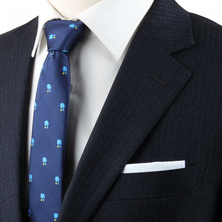 Close-up of an individual in a dark suit with a white dress shirt and a Pickle Ball Themed Tie adorned with small light blue objects, exuding refined charm. A white pocket square is visible in the suit jacket's breast pocket.