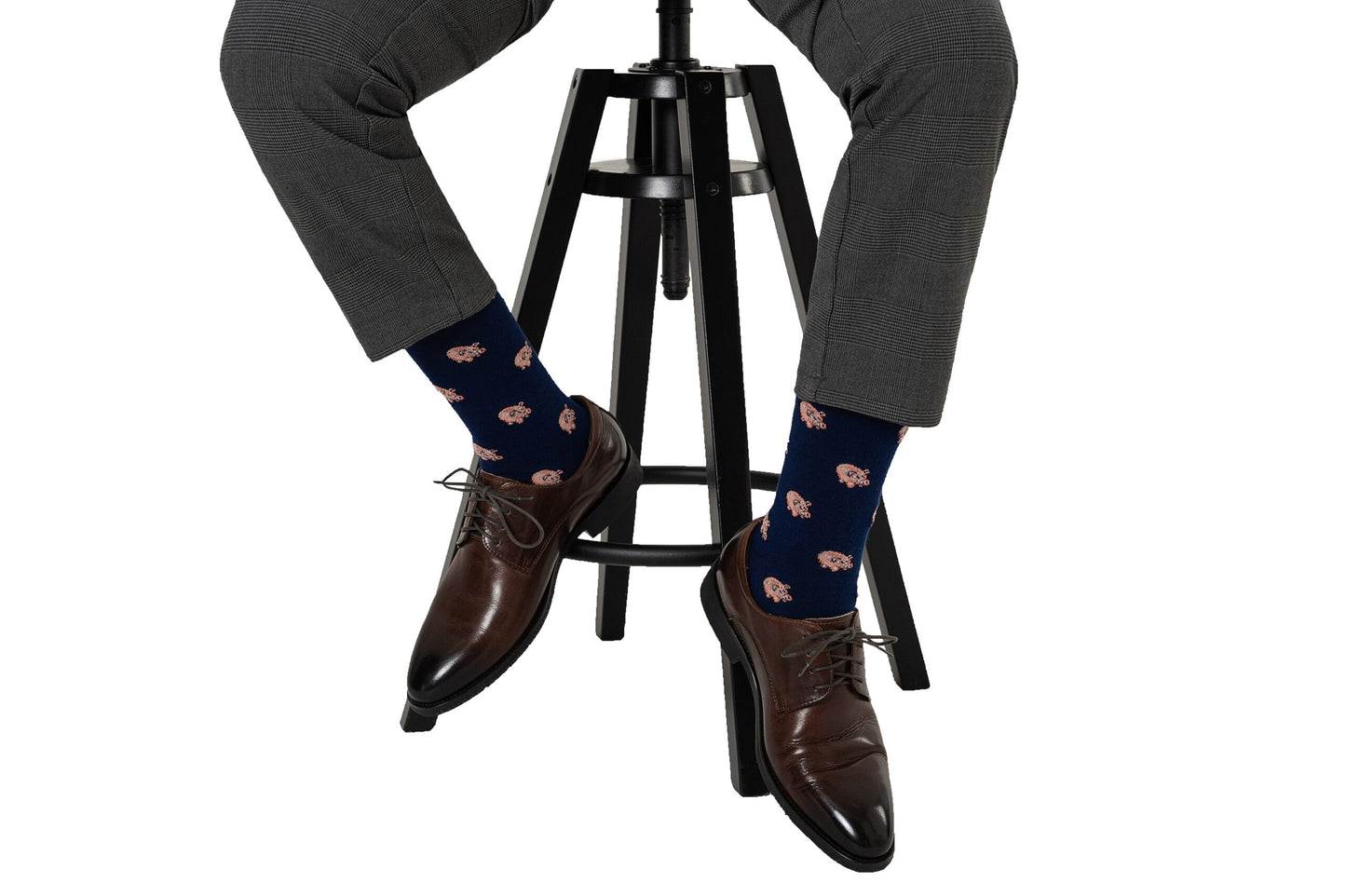 A person sitting comfortably on a stool, showcasing a fashion-forward look with grey trousers, stylish brown leather shoes, and dark blue Piggy Bank Socks adorned with a red pattern.