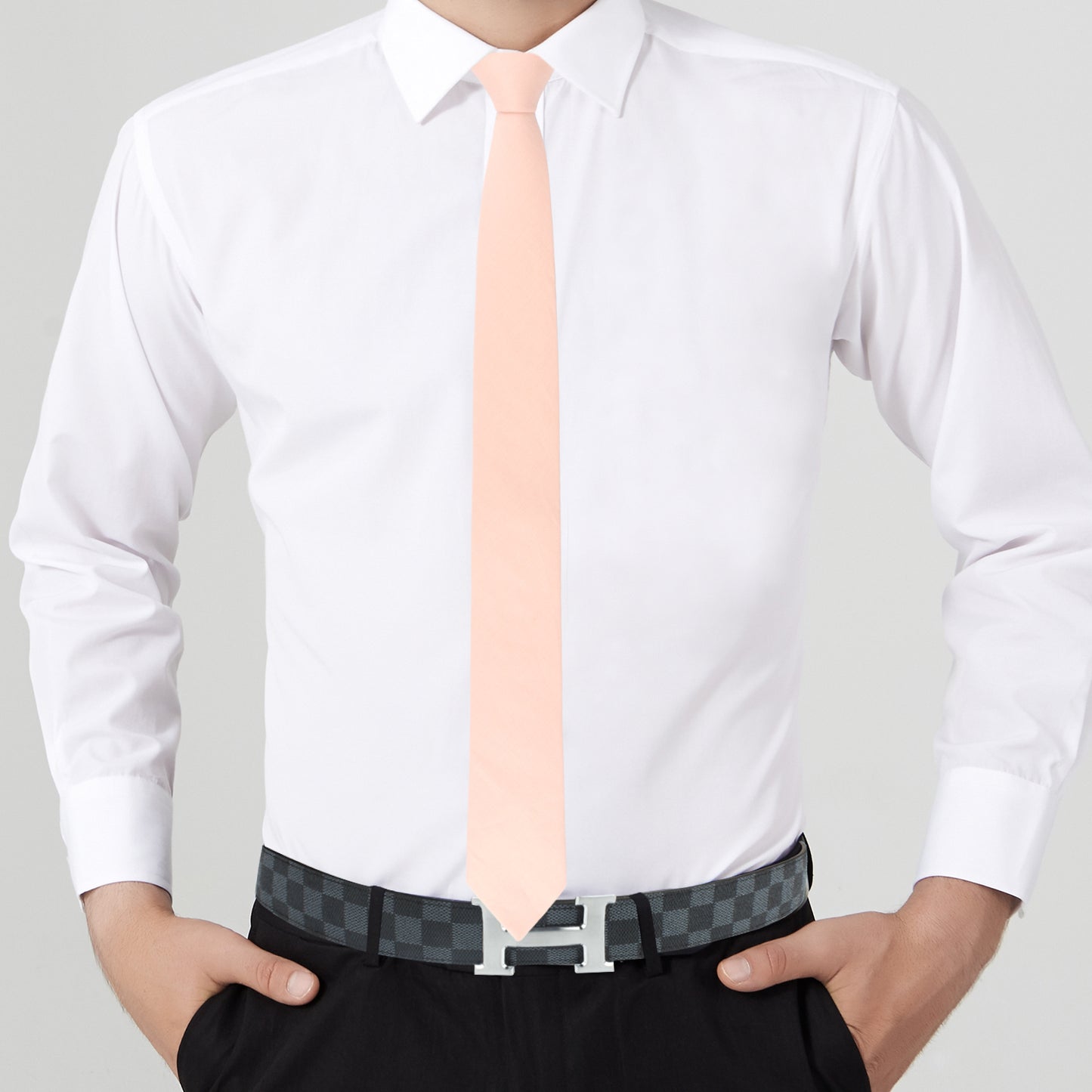 A person wearing a white dress shirt with a Baby Pink Skinny Tie and a checkered belt, woven with pastel threads, has hands in pockets.