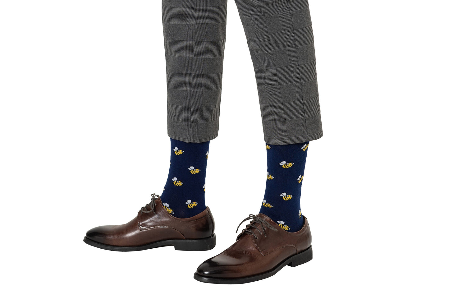 A person stands on a white background wearing Bee Socks, blue with a bee print, along with brown dress shoes and gray plaid trousers cropped above the ankle.