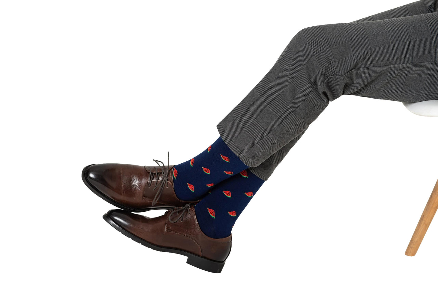A person wearing Watermelon Socks, brown dress shoes, and gray dress pants sits cross-legged on a white chair.