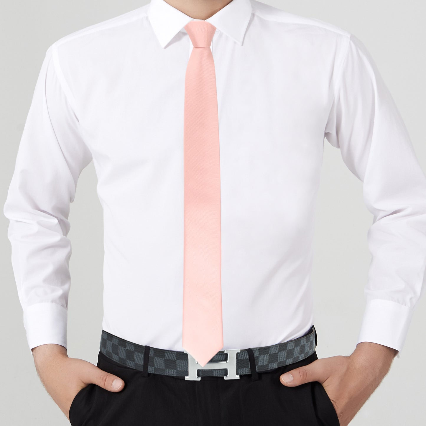 Person exuding modern elegance in a white dress shirt, sleek rose gold skinny necktie paired with a matching pocket square, black pants with a checkered belt, standing confidently with hands in pockets.