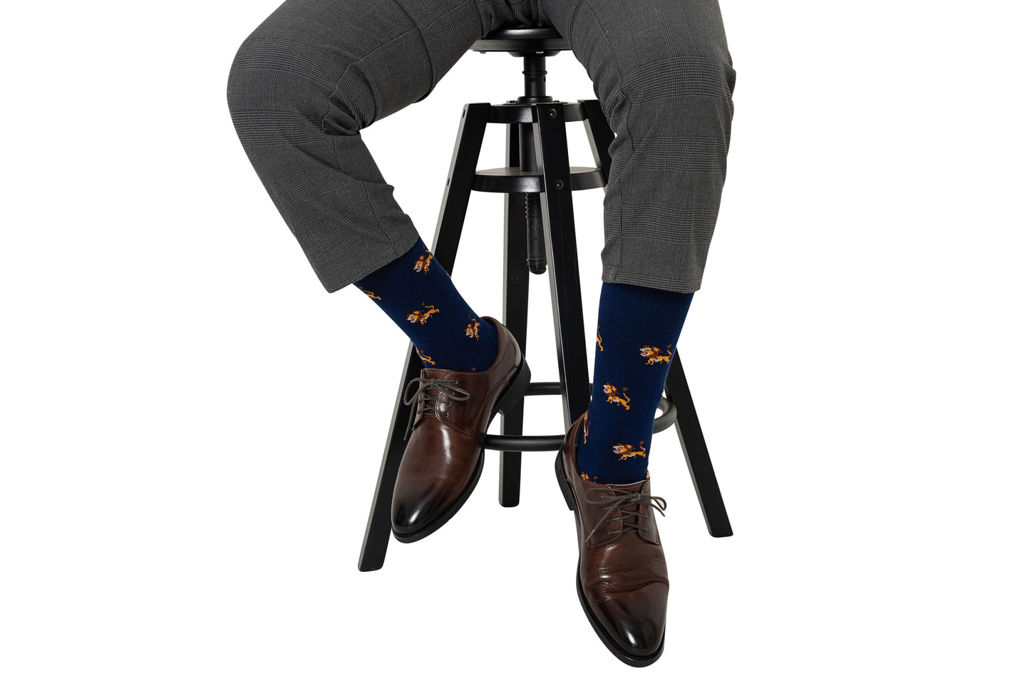 A person is seated on a black stool against a white background, wearing gray pants, brown dress shoes, and Lion Socks—navy blue socks featuring an orange lion pattern.