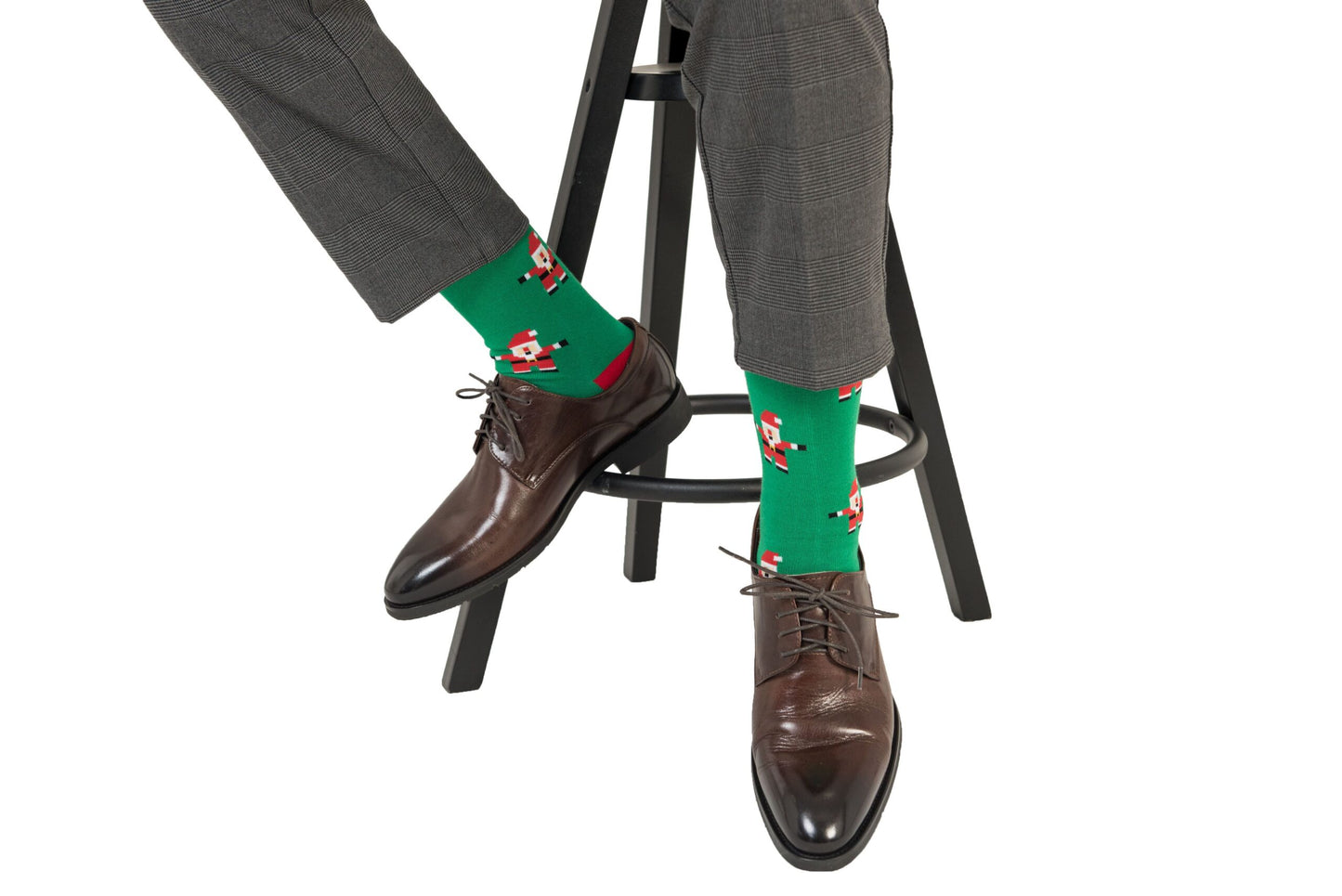 A person wearing gray pants, Santa Socks with a green background and Santa Claus print, and brown dress shoes is sitting on a black stool, showcasing their festive feet.