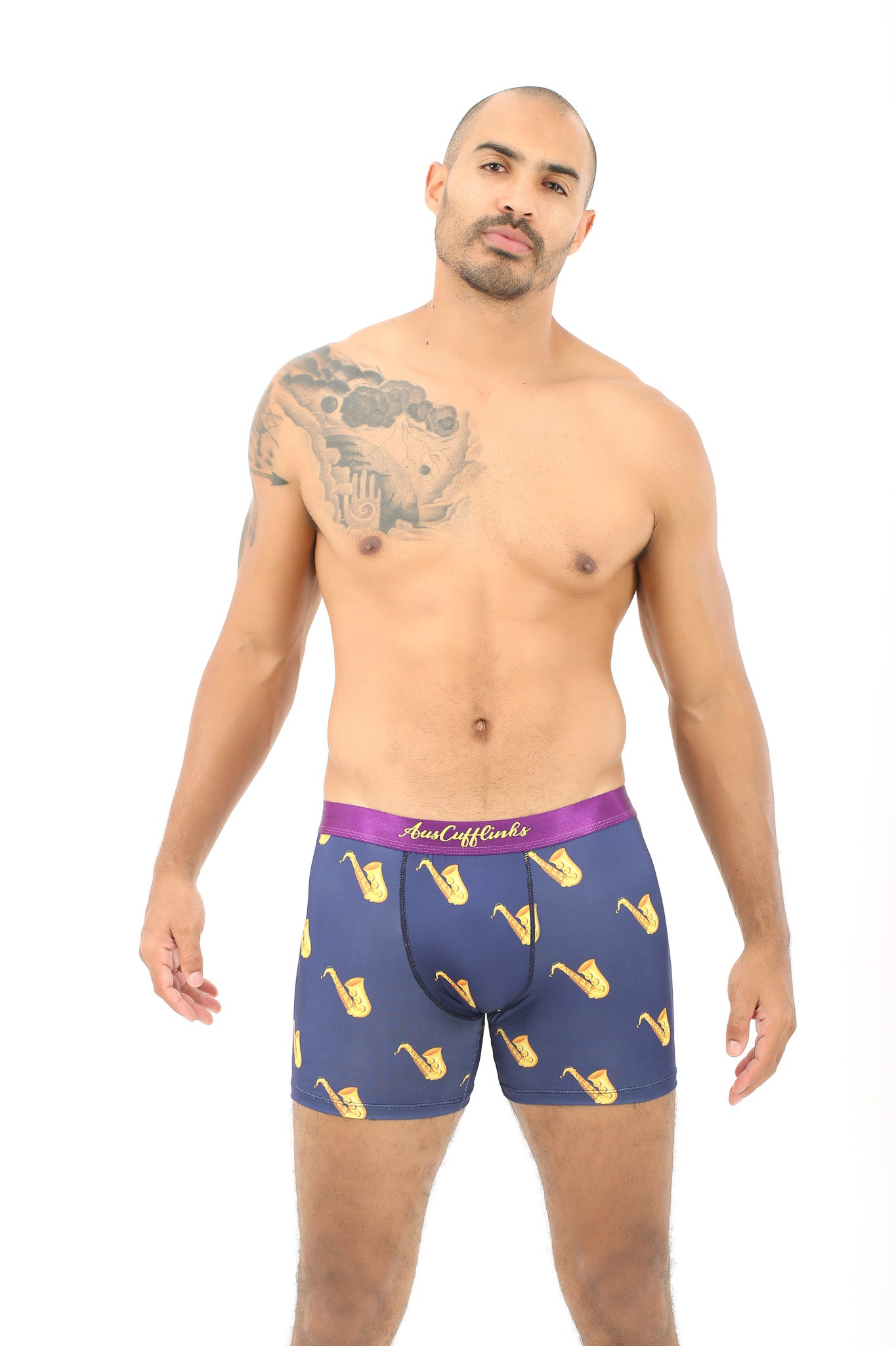 A shirtless man with a visible arm tattoo models the Saxophone Underwear – Fun Gift for Music Lovers: navy blue boxer briefs with yellow saxophone prints and a purple waistband, standing against a plain white background.