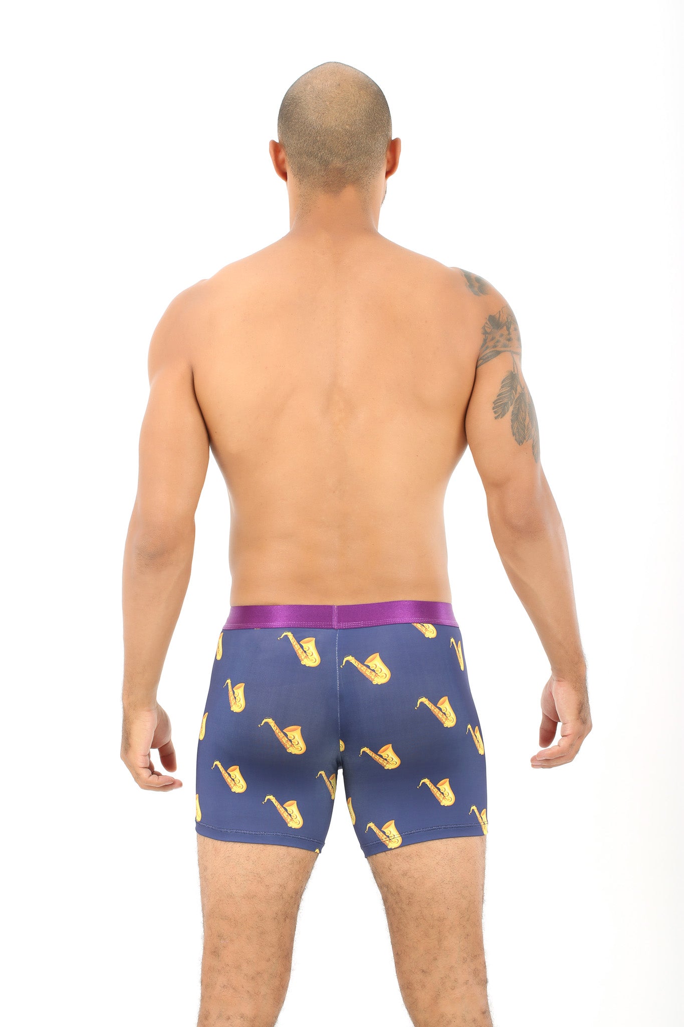 A man with a shaved head and arm tattoo stands facing away, wearing Saxophone Underwear – Fun Gift for Music Lovers, featuring a blue jazz-inspired design with yellow saxophones and a purple waistband.