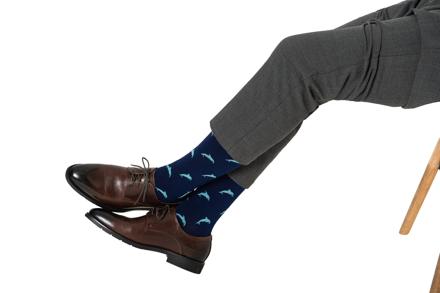 Someone is sitting cross-legged on a wooden chair, wearing gray suit pants, brown dress shoes, and Dolphin Socks—ocean-themed novelty socks featuring dolphin patterns.
