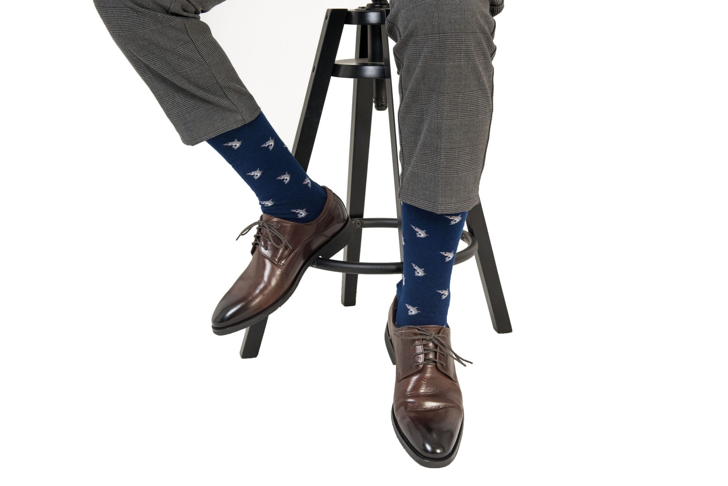 A seated person in gray plaid pants and dark brown leather shoes, crossing their legs with an air of style, is wearing Shark Socks in navy blue, featuring a small white pattern reminiscent of the deep sea.