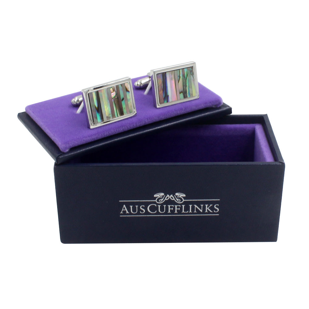A pair of Shell Stone Cufflinks with abalone inlay, showcasing nature's finesse, rests elegantly on a purple velvet stand inside a sleek black box labeled "AusCufflinks.