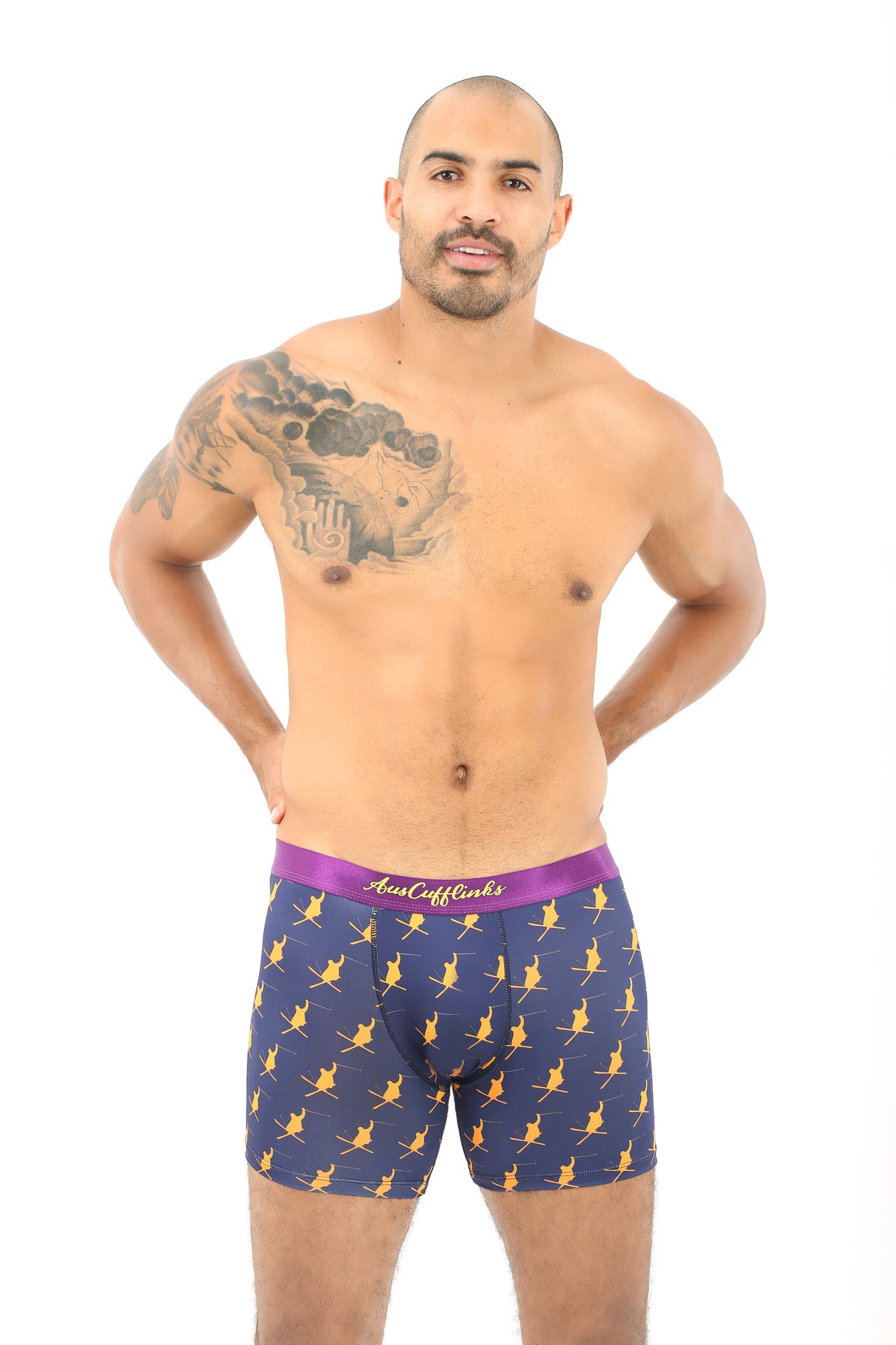 A man with a tattoo wearing Ski Underwear stands against a white background. The patterned boxer briefs, ideal for winter sports enthusiasts, feature a purple waistband and offer moisture-wicking properties for enhanced comfort.