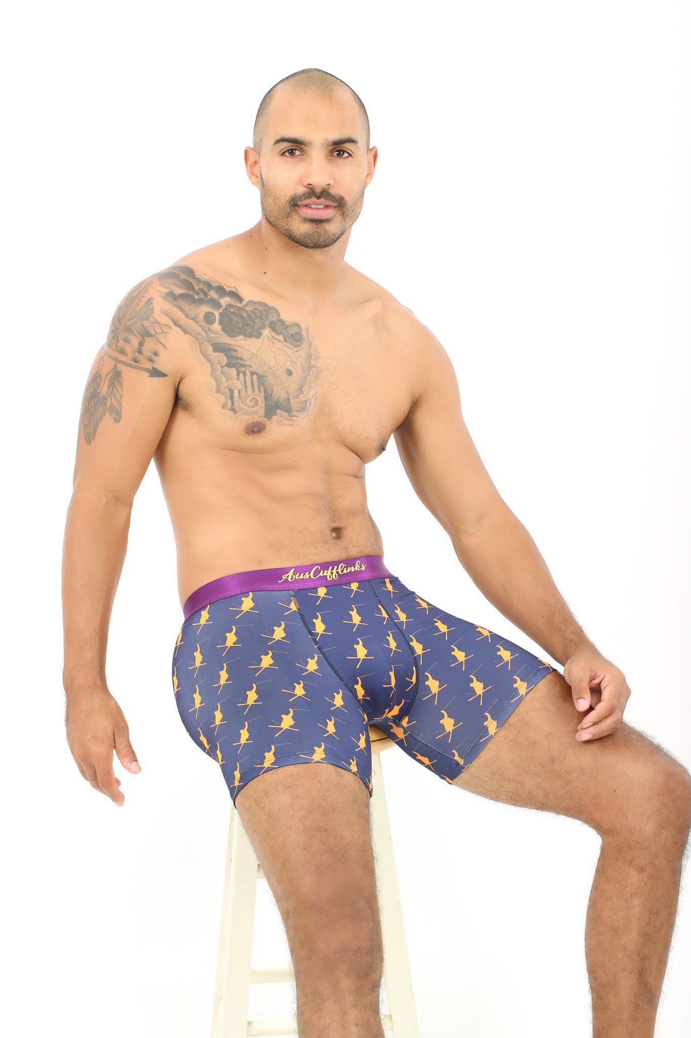 A shirtless man with an arm tattoo sits on a stool, showcasing the Ski Underwear: blue boxer briefs with a yellow pattern. These moisture-wicking boxers are perfect for staying cool during winter sports.
