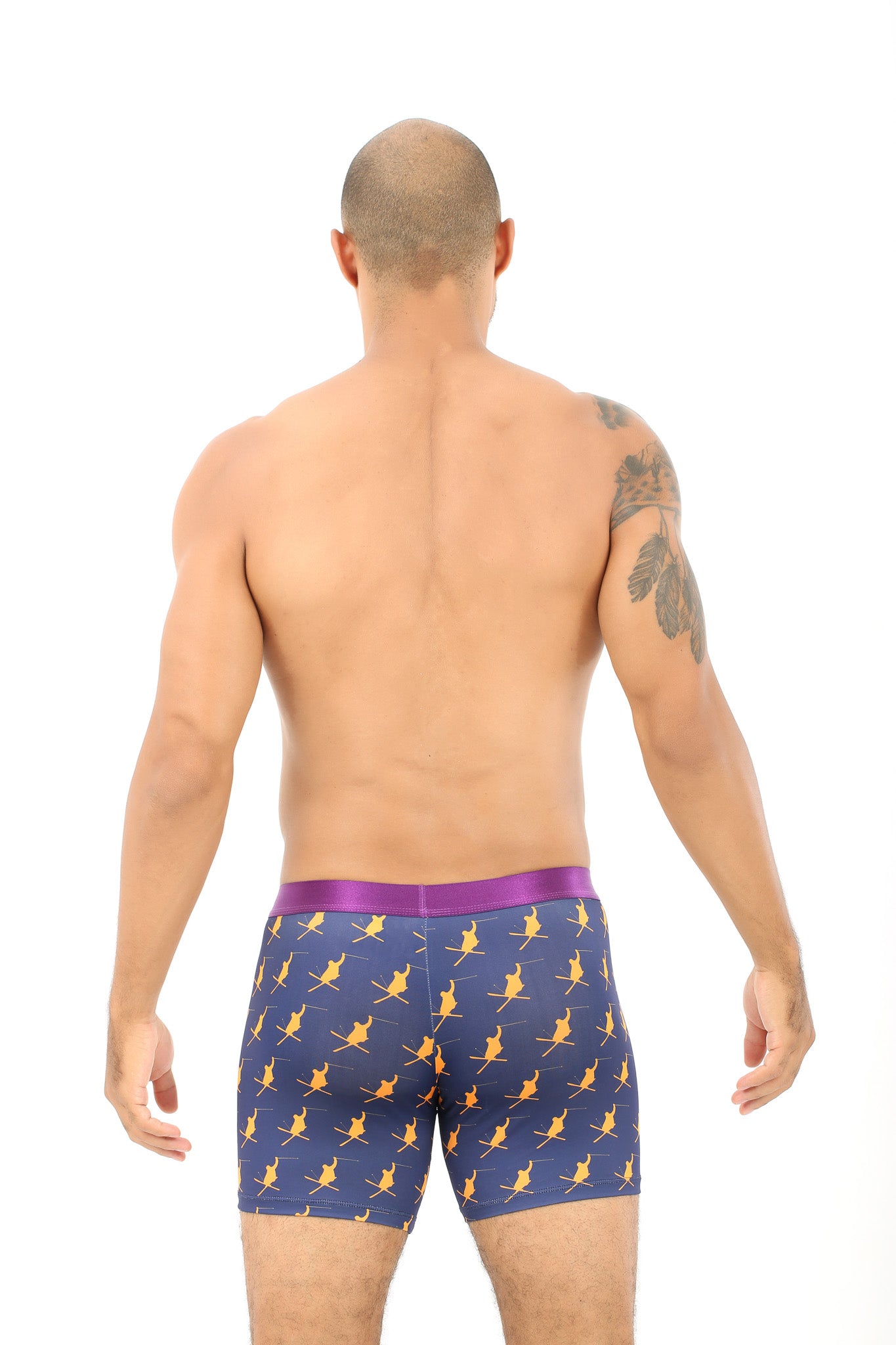 A man with a shaved head and tattooed arm is wearing Ski Underwear, featuring navy boxer briefs with yellow designs made from moisture-wicking fabric, viewed from the back against a plain white background.