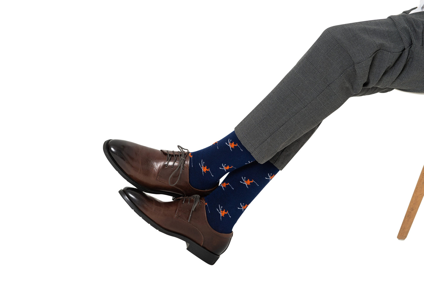 A person sits cross-legged on a chair against a white background, wearing grey dress pants, brown dress shoes, and blue Ski Socks featuring a skier print.