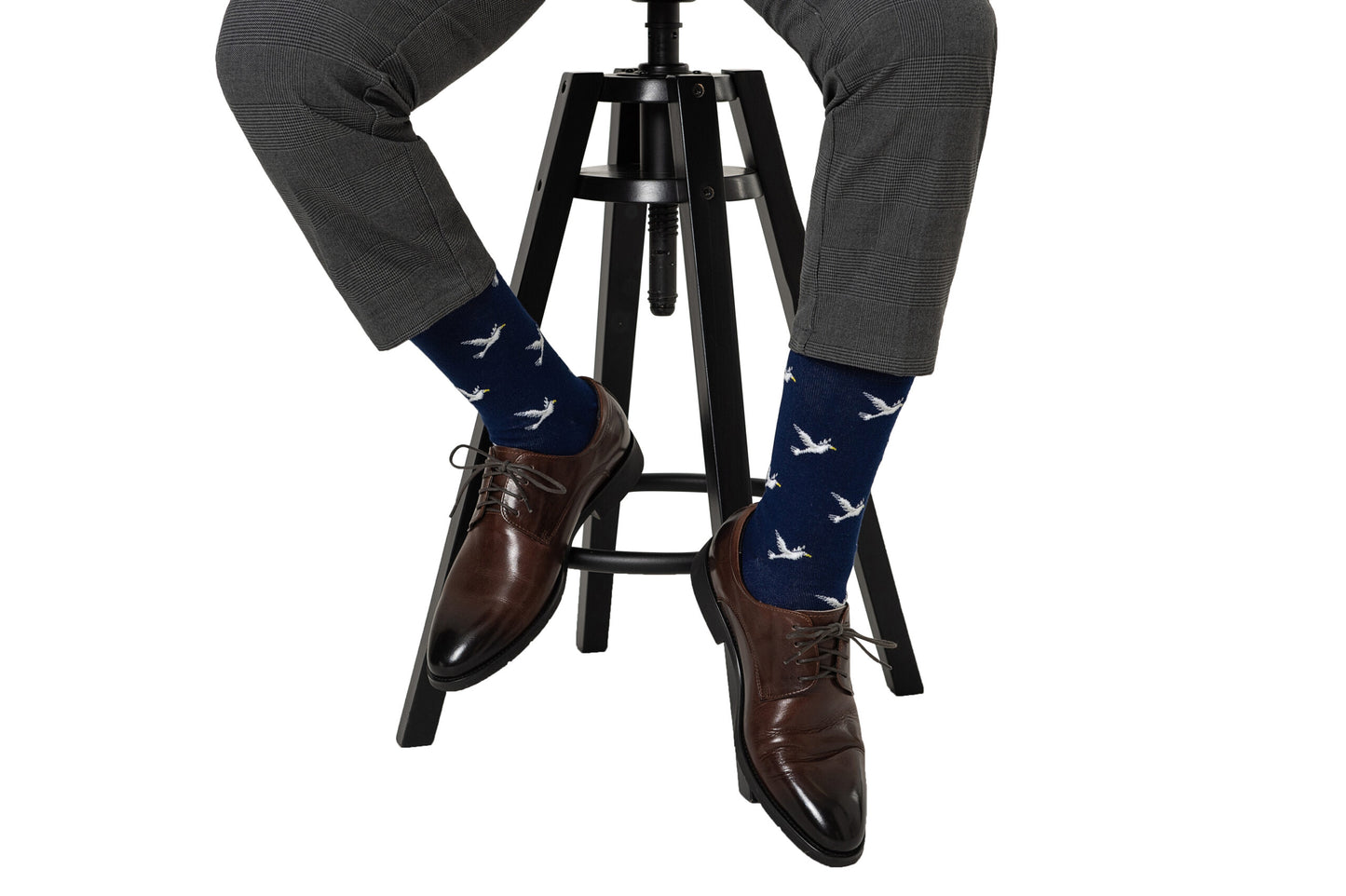 A person in gray dress pants and brown shoes sits on a stool, wearing Dove Socks—navy blue socks featuring a white bird pattern.