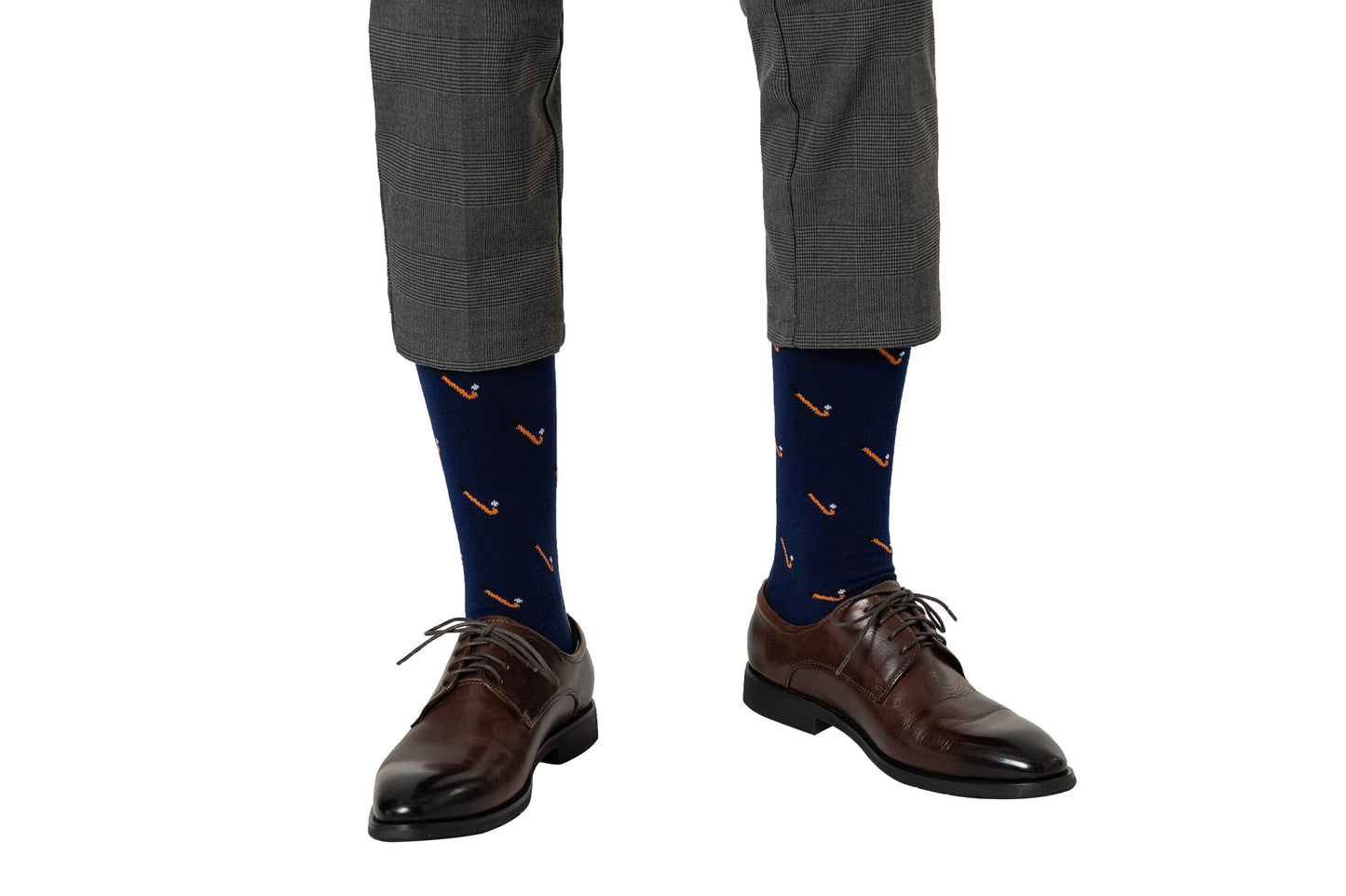 A person is wearing grey plaid pants, brown dress shoes, and navy blue Field Hockey Socks featuring orange pipe patterns, set against a white background.