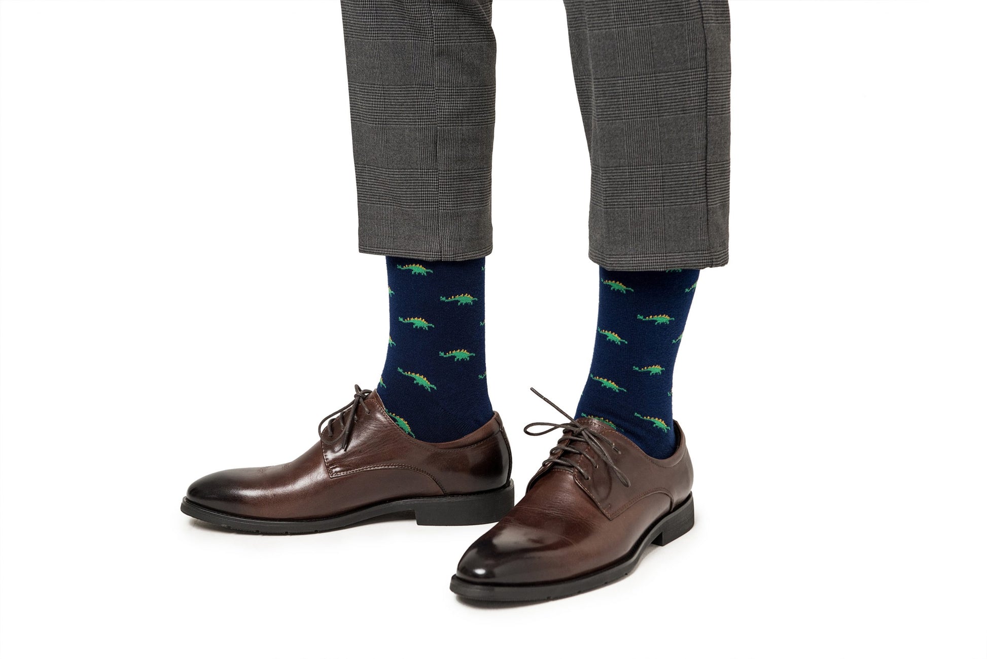 A person standing in brown dress shoes and Stegosaurus Socks with whimsical green alligator patterns, against a white background.