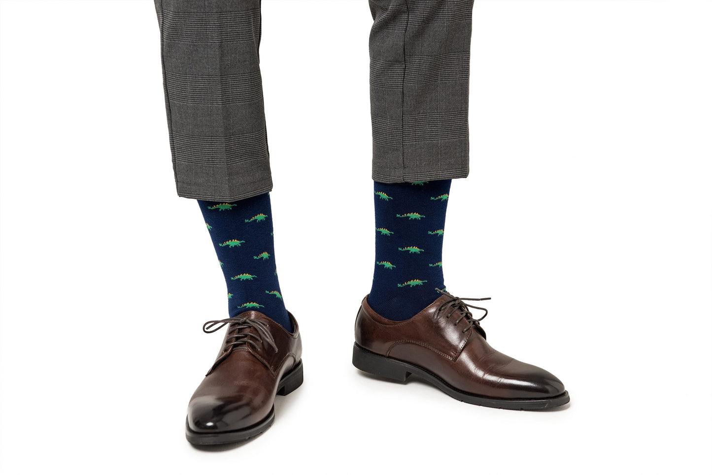 A person wearing brown dress shoes and Stegosaurus Socks adorned with whimsical green dinosaur patterns, paired with grey trousers.