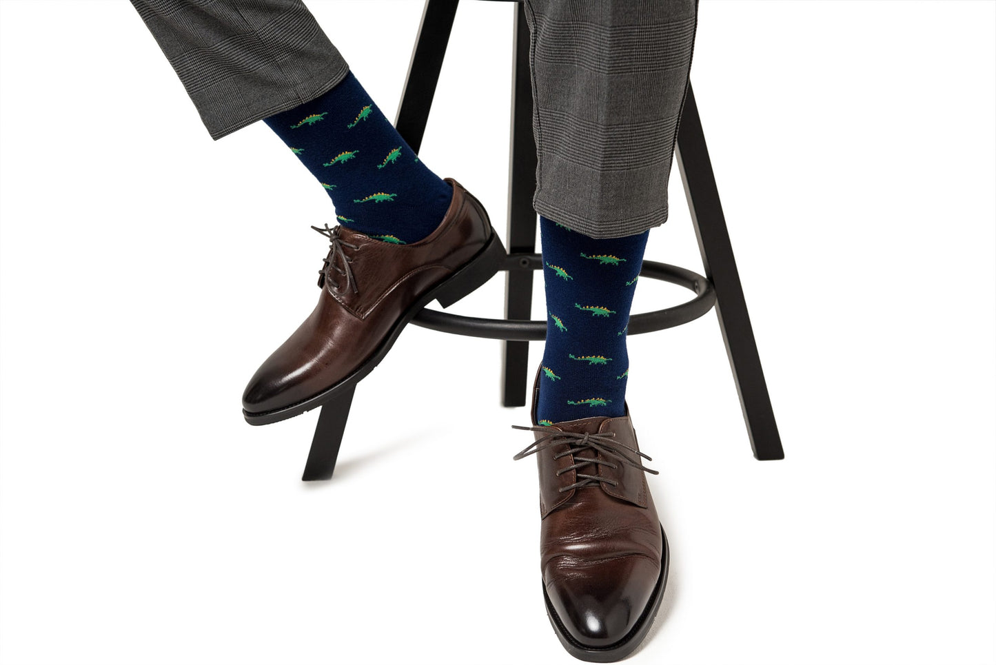 Person wearing grey trousers, Stegosaurus socks, and brown leather shoes sitting on a black stool.