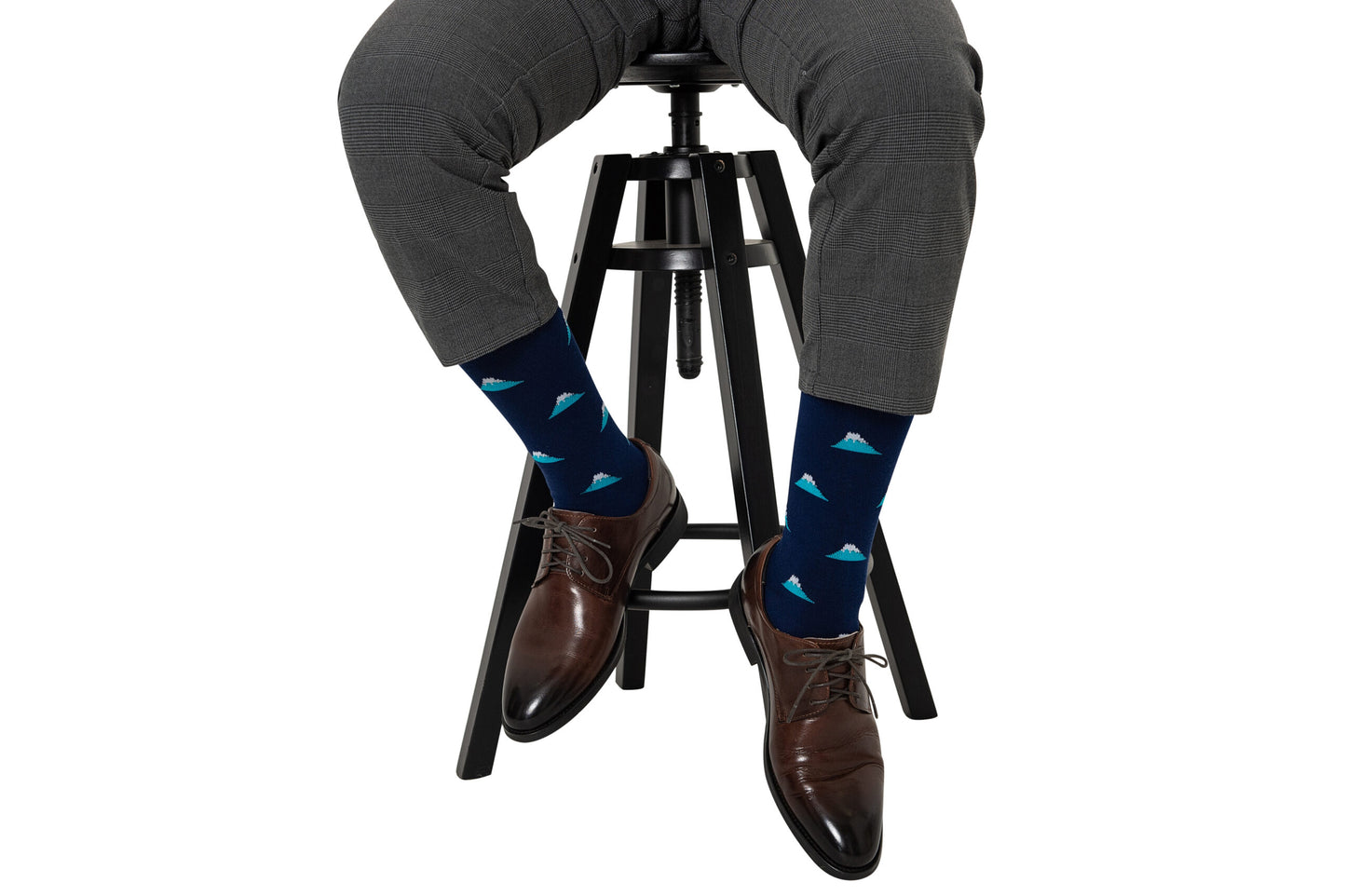 Man in gray pants and brown dress shoes sitting on a black barstool, wearing Mountain Socks with blue triangular patterns that resemble peaks and valleys.
