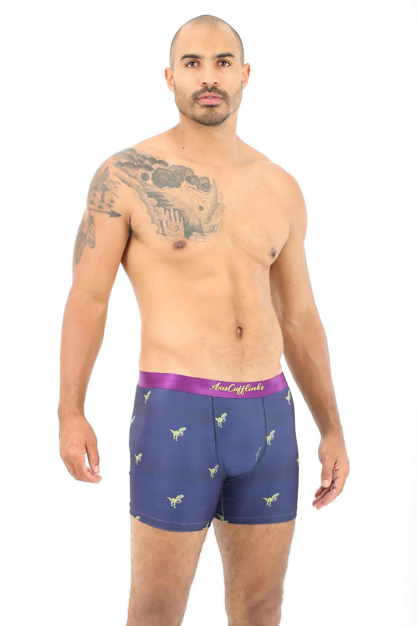 A man with a shaved head and tattoos wears T-Rex Underwear, featuring navy blue boxer briefs with a purple waistband and small green dinosaur patterns. Standing against a white backdrop, he exudes roaring comfort with his prehistoric-themed style.
