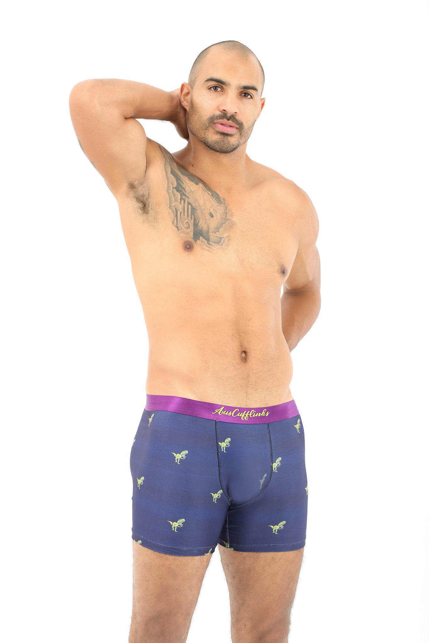 A tattooed man poses shirtless in T-Rex Underwear, featuring purple and blue boxer briefs with small green dinosaur designs, against a plain white background.