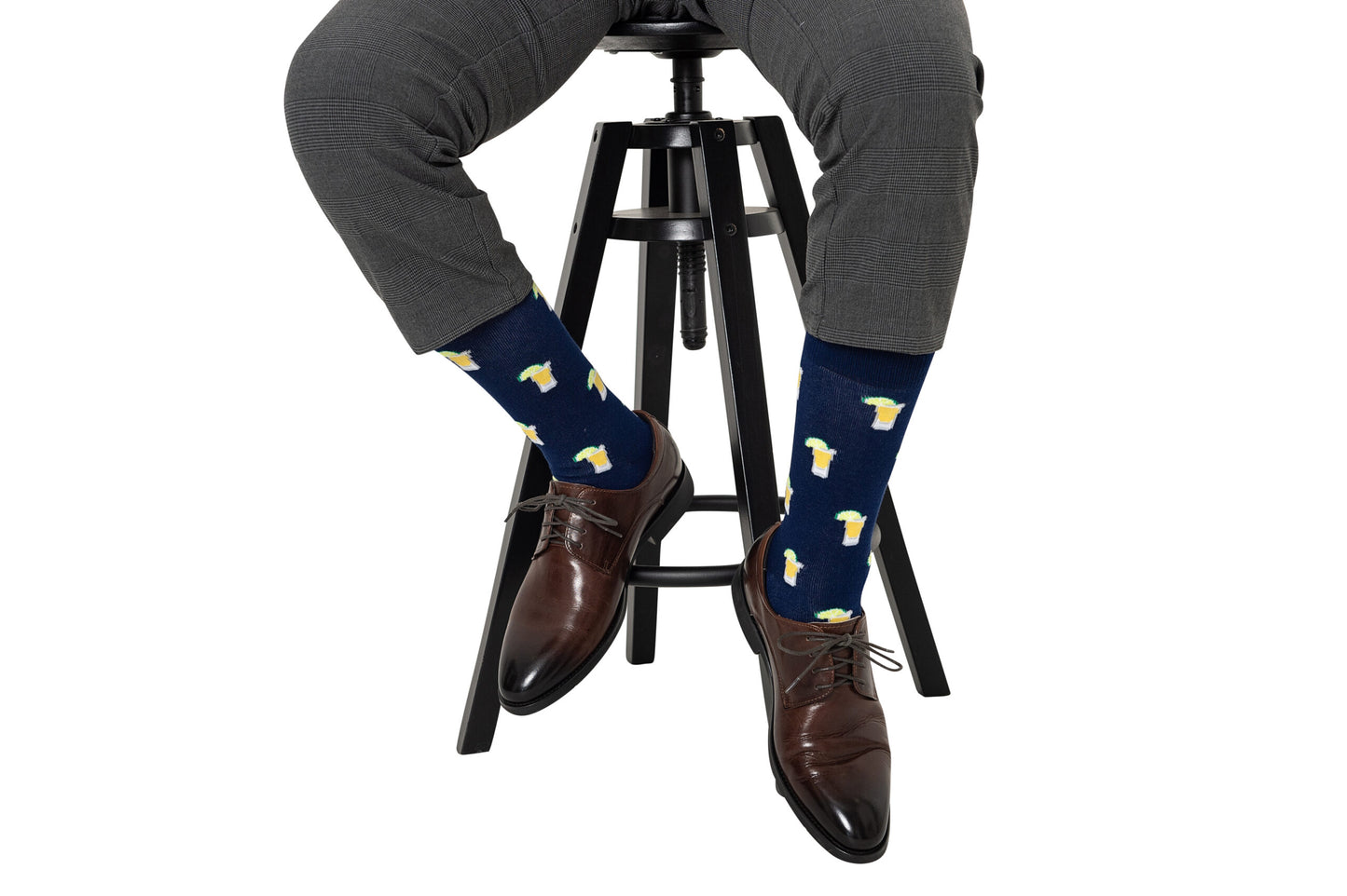 Wearing Tequila Socks featuring a beer mug pattern, a person in gray pants and brown dress shoes sits on a black stoolâideal party socks for any tequila fan.
