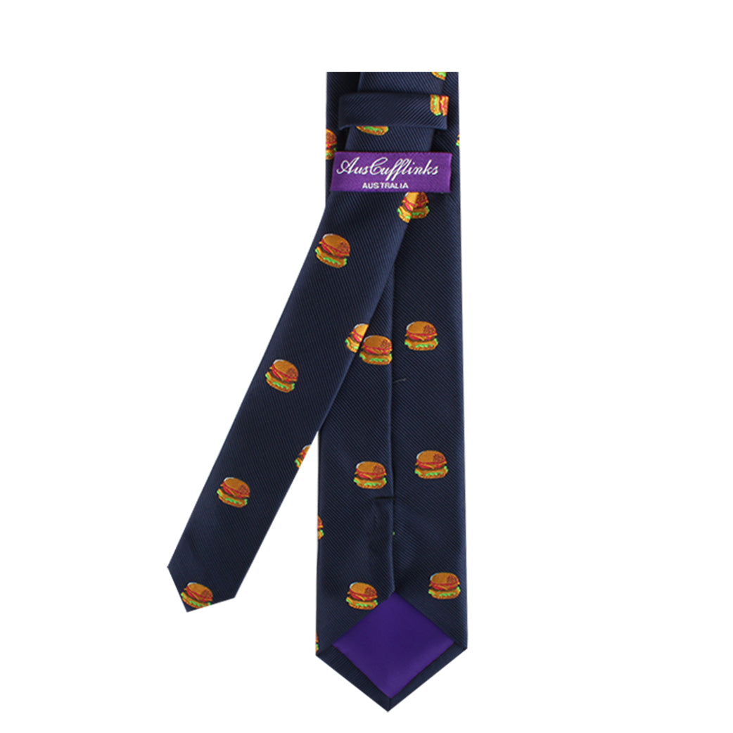 The Burger Skinny Tie, featuring a bold navy blue hue and an eye-catching pattern of small hamburger images, is a stylish statement piece. It includes a purple label with striking white text.
