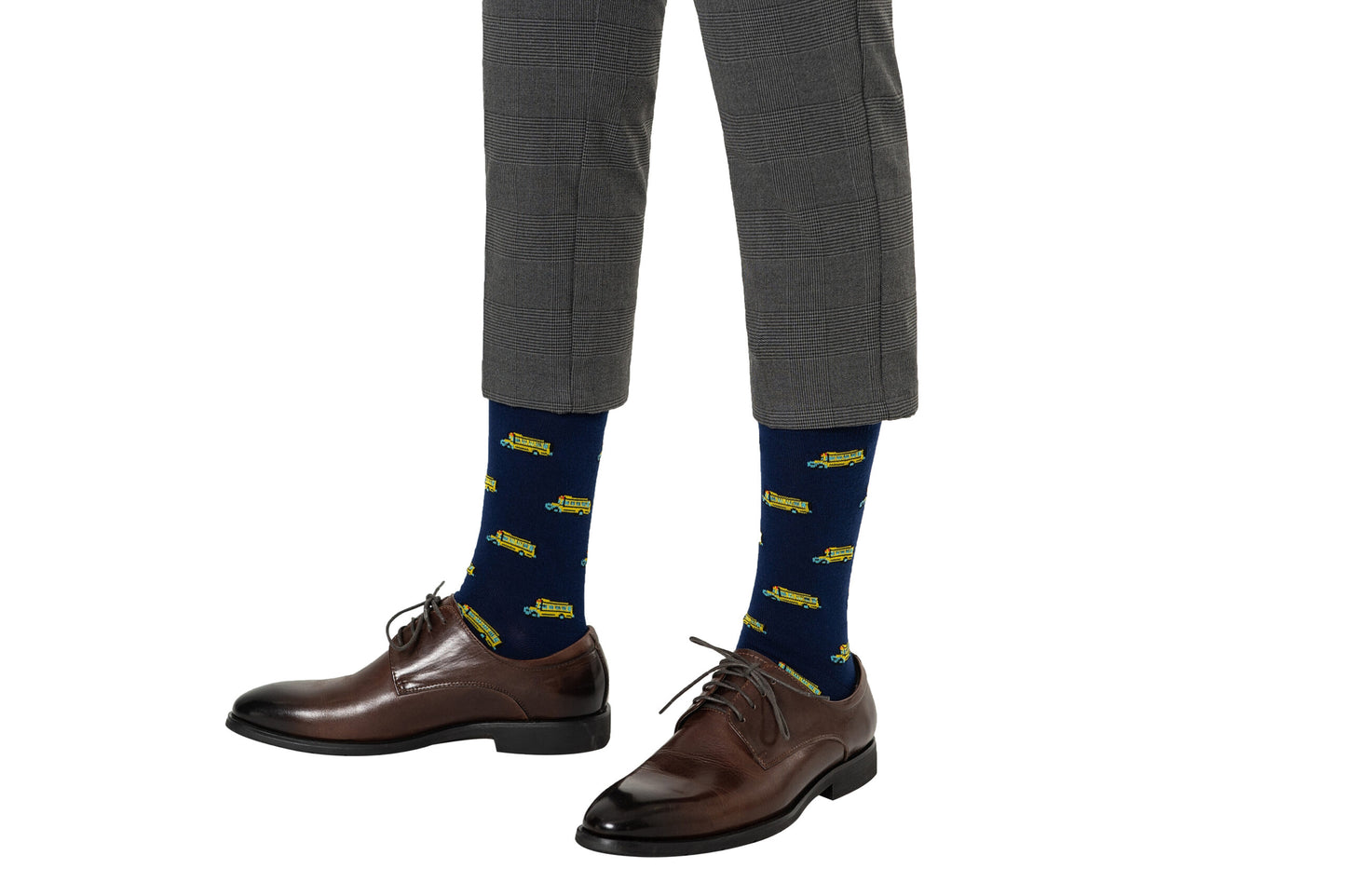 Close-up of a person wearing dark brown dress shoes, Transportation Socks featuring green and yellow patterns on a blue background, and grey checkered pants that evoke a sense of nostalgia. The person's feet are slightly apart, and the pants are cropped above the ankles, capturing a timeless roll in their step.