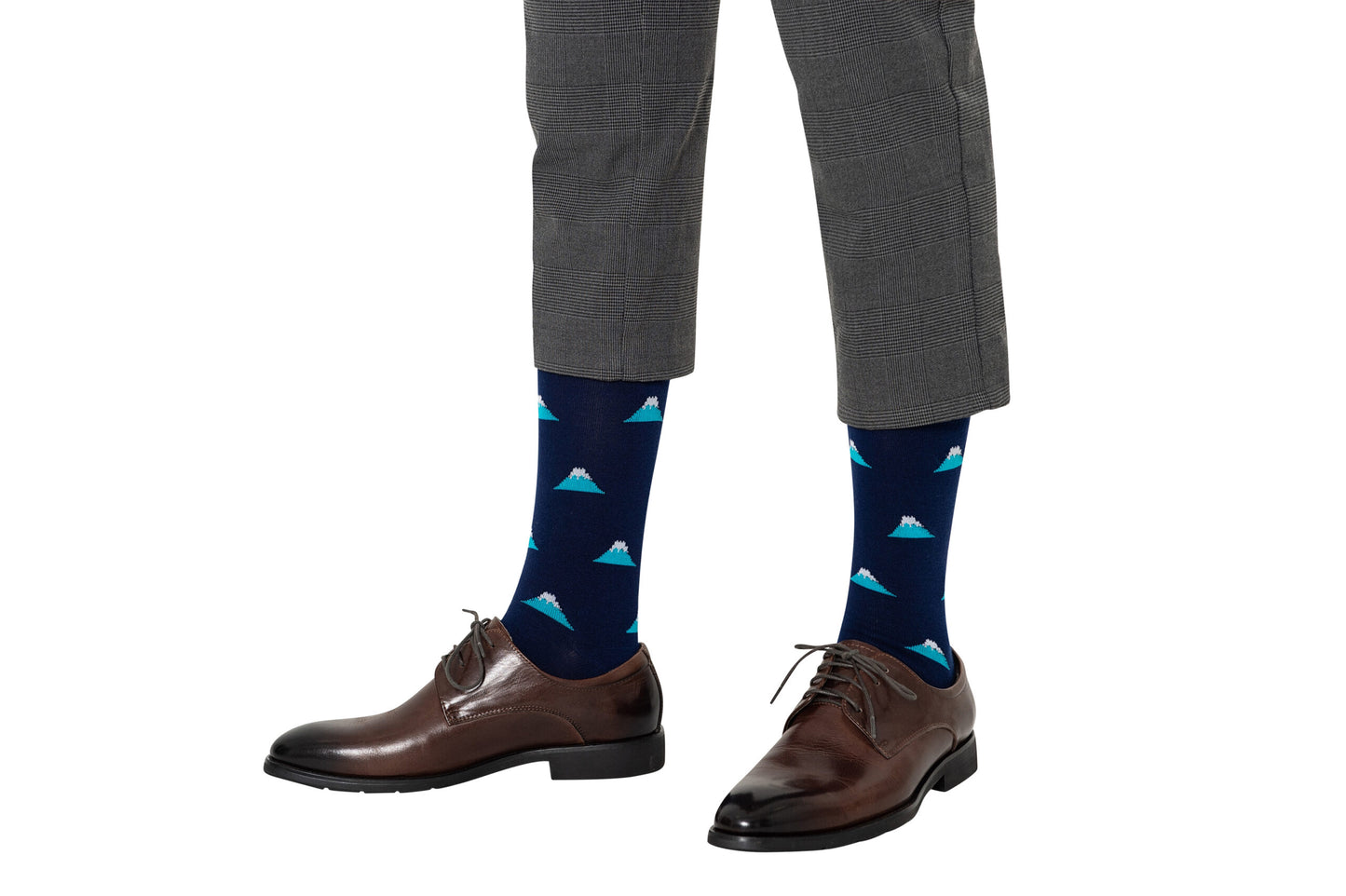 A close-up captures a person exuding style in gray pants and dark dress shoes, while wearing Mountain Socks—blue with a mountain pattern—that peak above the calves beneath slightly cropped pants.