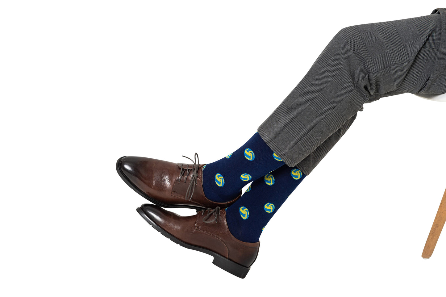A person in gray trousers and brown shoes sits with legs crossed, wearing Volleyball Socks—navy blue socks featuring a green and yellow volleyball pattern—a stylish choice for anyone seeking a unique gift.