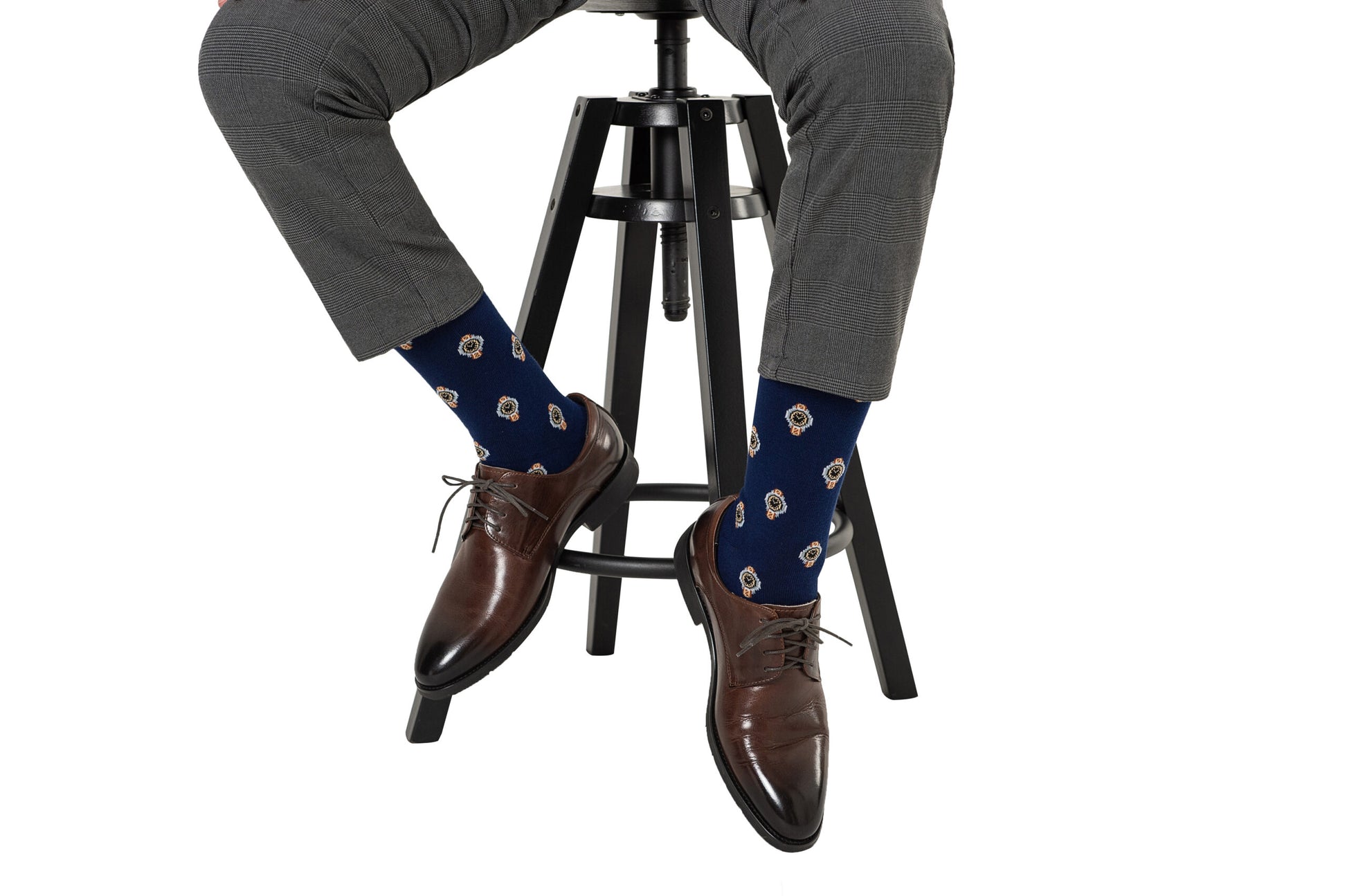 A person seated wearing brown leather shoes and blue Watch Socks with owl designs, perched on a black stool, showing only the lower half of their body, exudes timeless elegance.