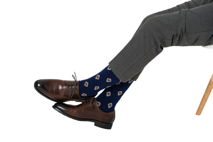 A person wearing dark brown leather dress shoes and blue Watch Socks with a timeless floral pattern, seated with legs crossed over a white background.