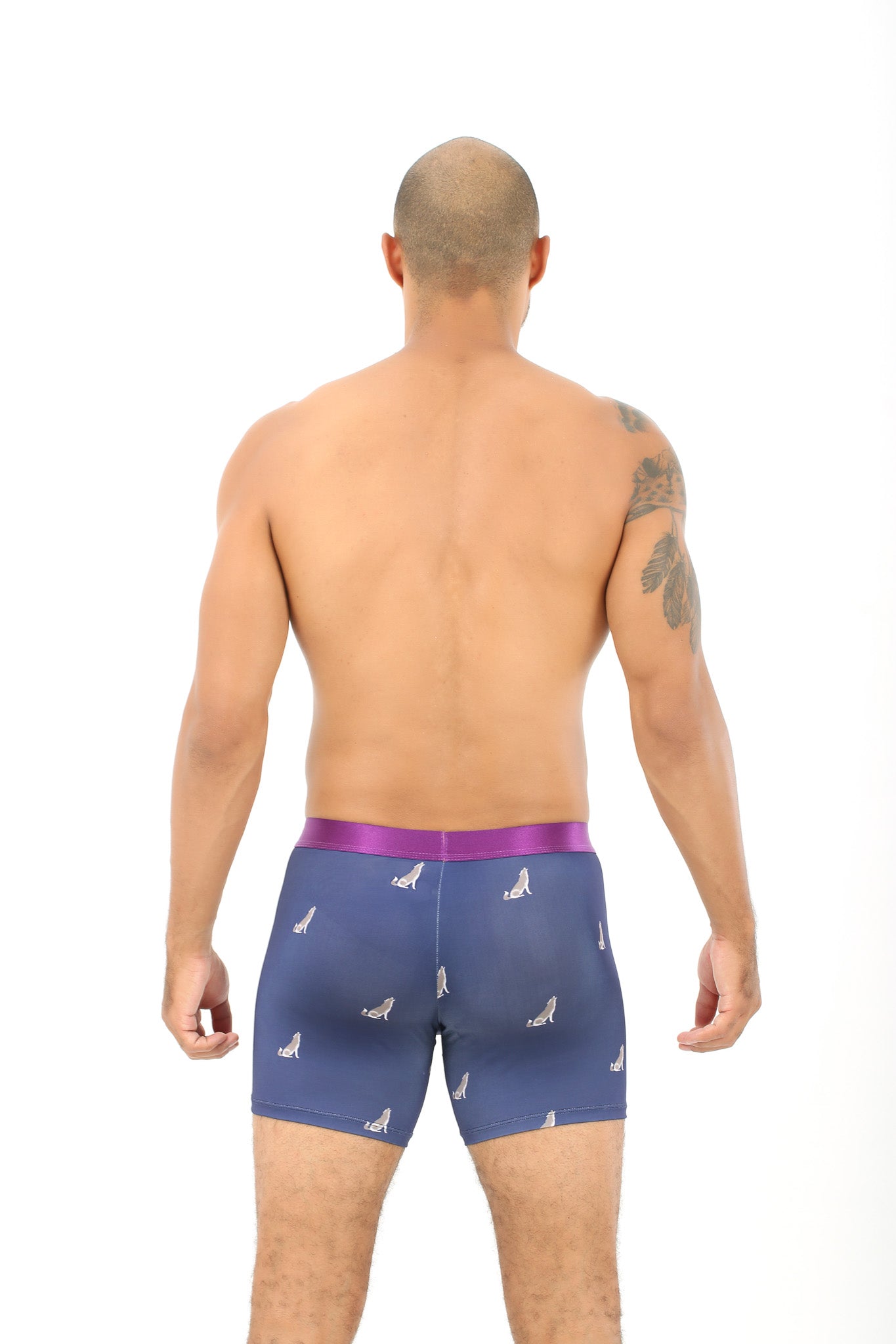 A person with a tattoo stands with their back to the camera, wearing Wolf Underwear—a pair of blue boxer briefs showcasing a wolf print and a purple waistband.