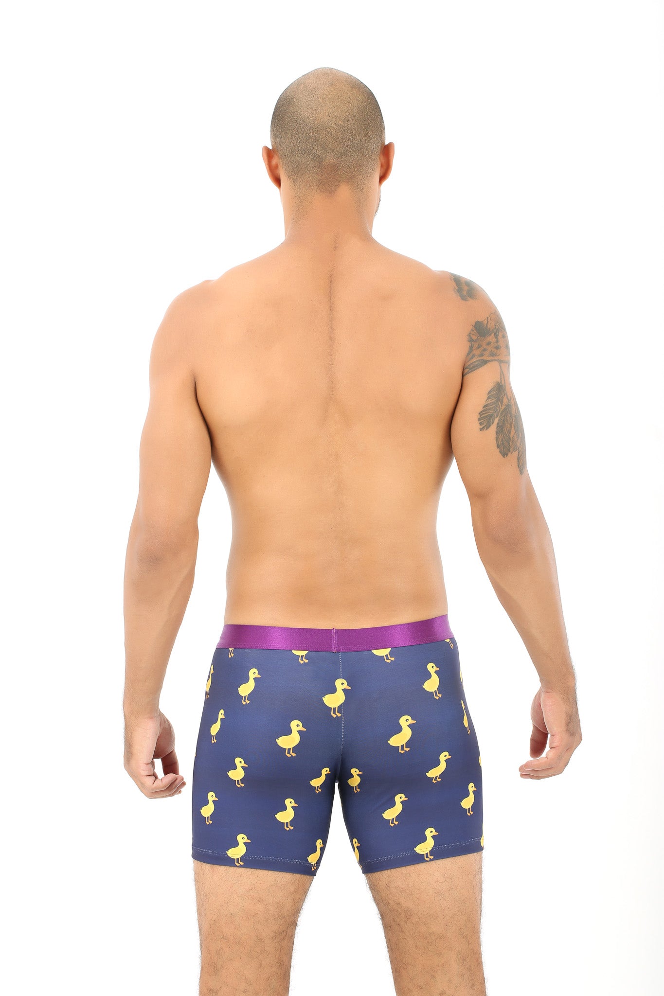 A man faces away, displaying his shaved head and upper body. He wears Duck Underwear for Men – Gift for Animal & Bird Lovers: blue boxer briefs featuring a yellow duck print and a purple waistband.