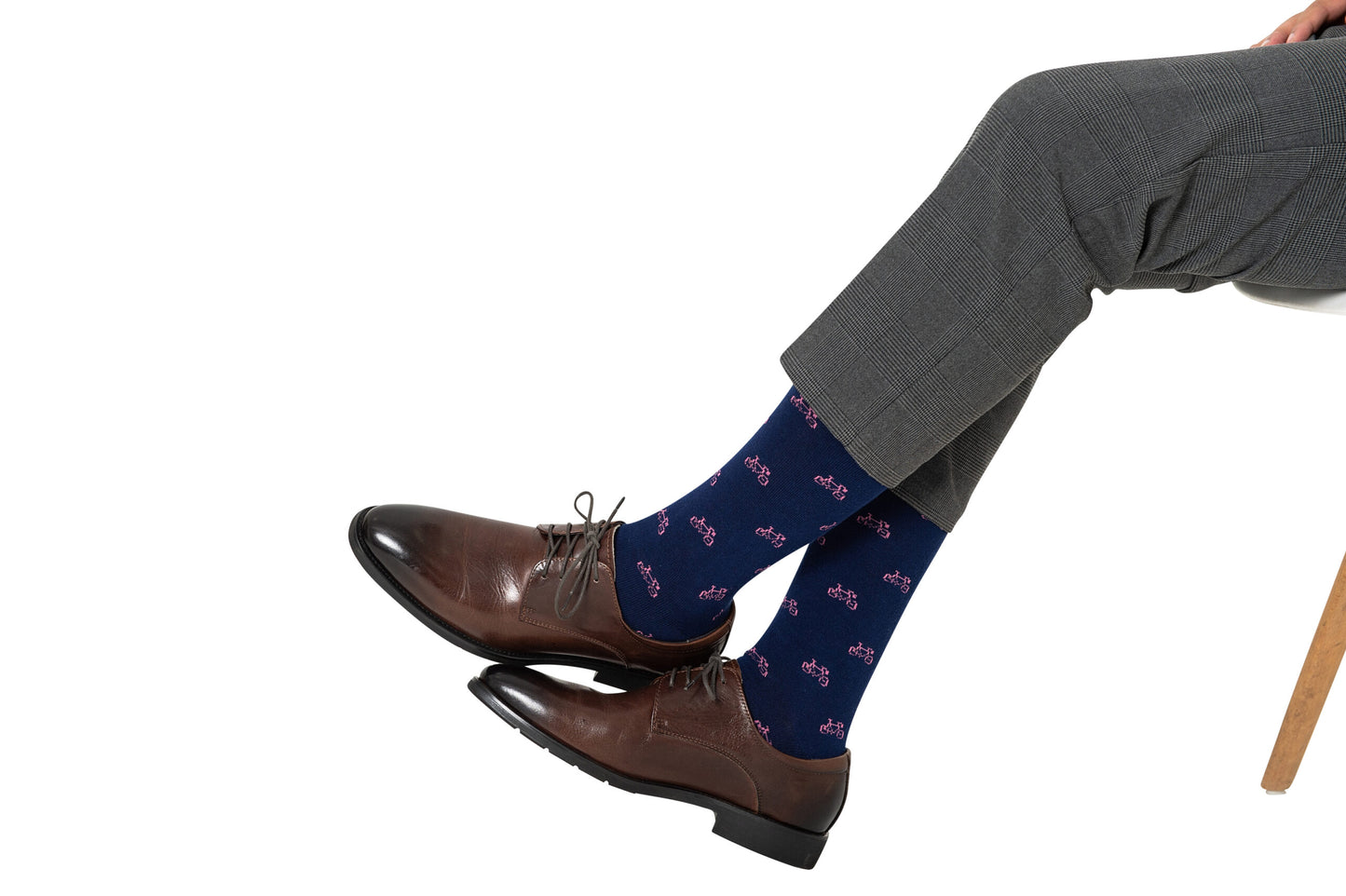A person sits comfortably on a chair, wearing gray pants, dark brown shoes, and Pink Bike Socks with bicycle patterns, perfect for those who love to pedal in style.