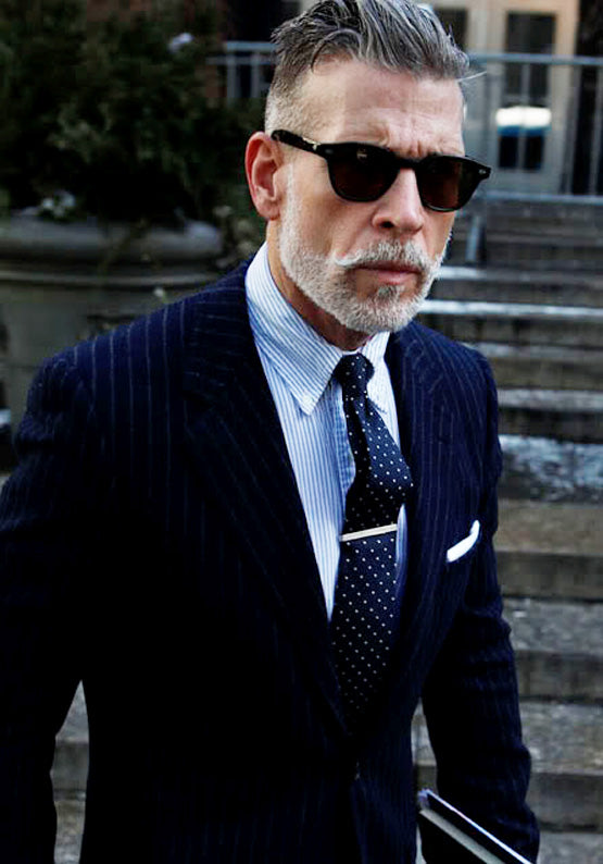 A man with grey hair and a beard, wearing a pinstripe suit, dotted tie, polished silver tie pin, and sunglasses, holds a book while standing outdoors near steps.