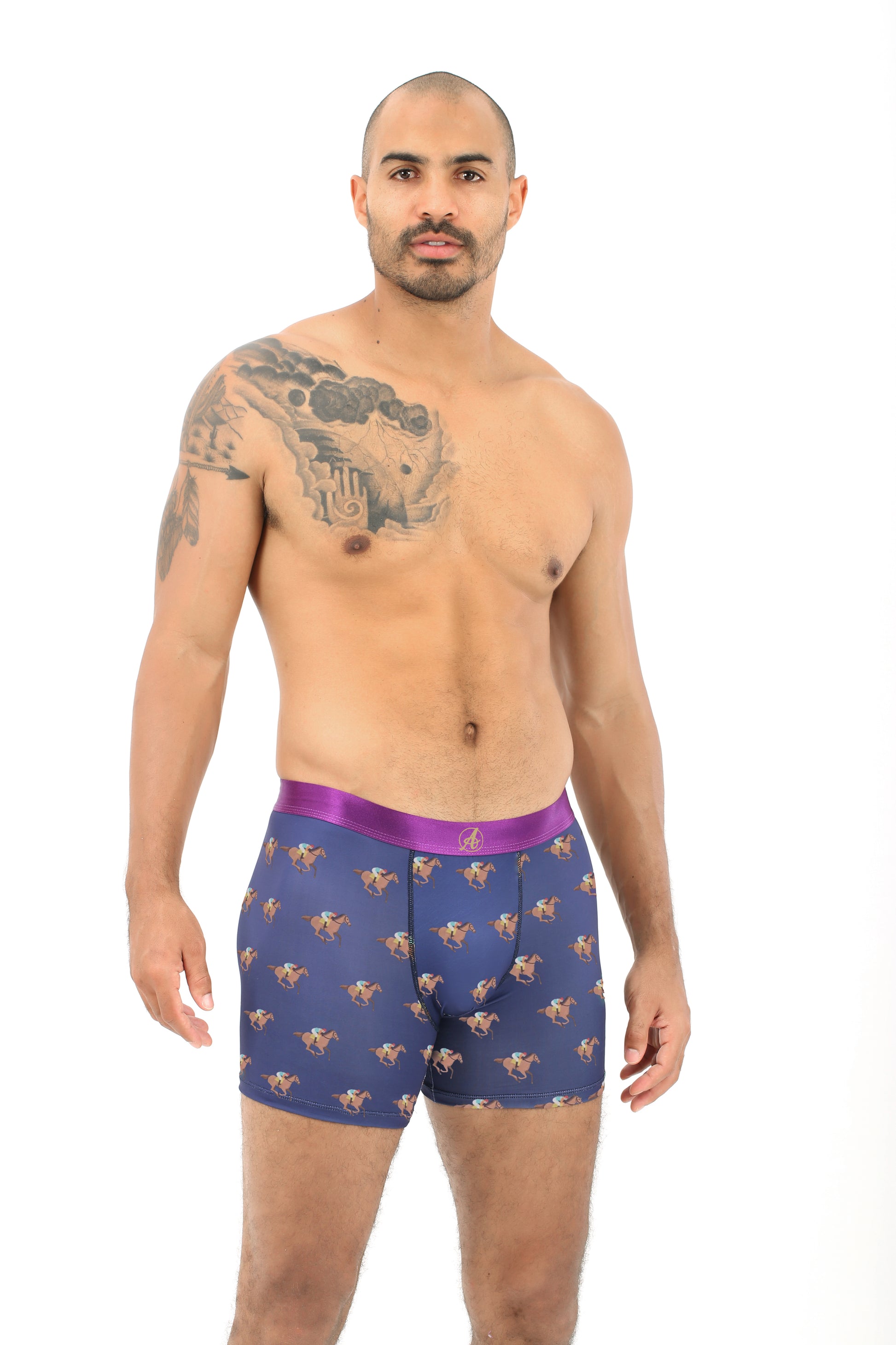 A man with a shaved head and tattooed arm stands against a white background, wearing blue Horse Racing Underwear boxer briefs featuring a jockey print, made from breathable poly-spandex and finished with a purple waistband.